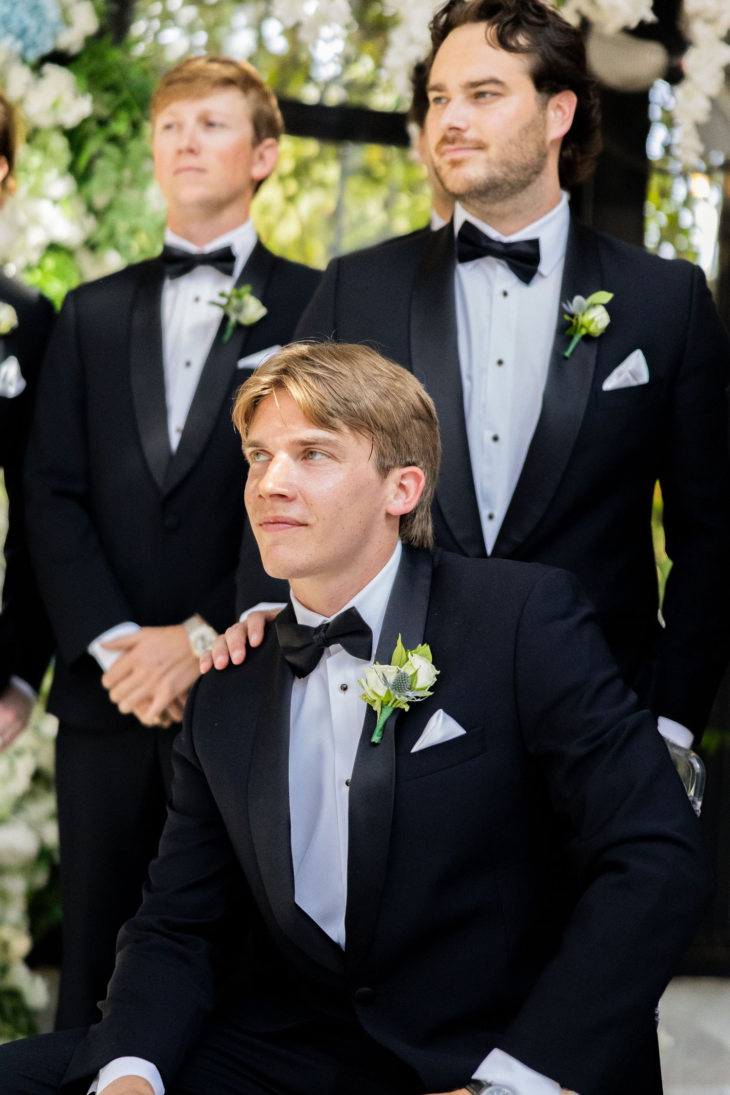 Groomsmen in black tuxedos with bow ties and boutonnieres pose together at an elegant outdoor wedding ceremony.