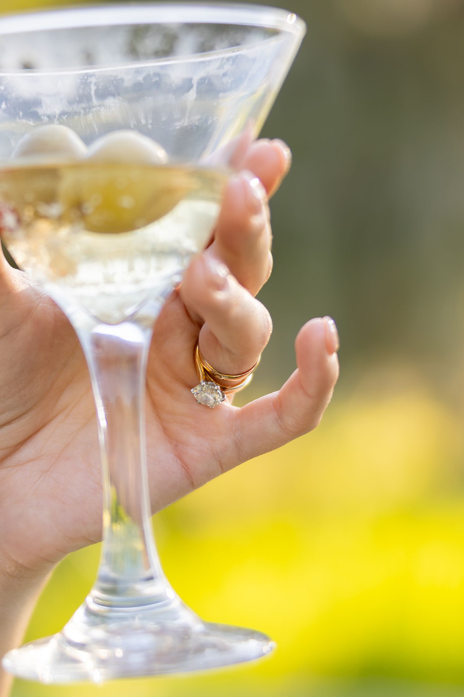 Hand with diamond engagement ring holding a martini glass with olives outdoors in natural light.