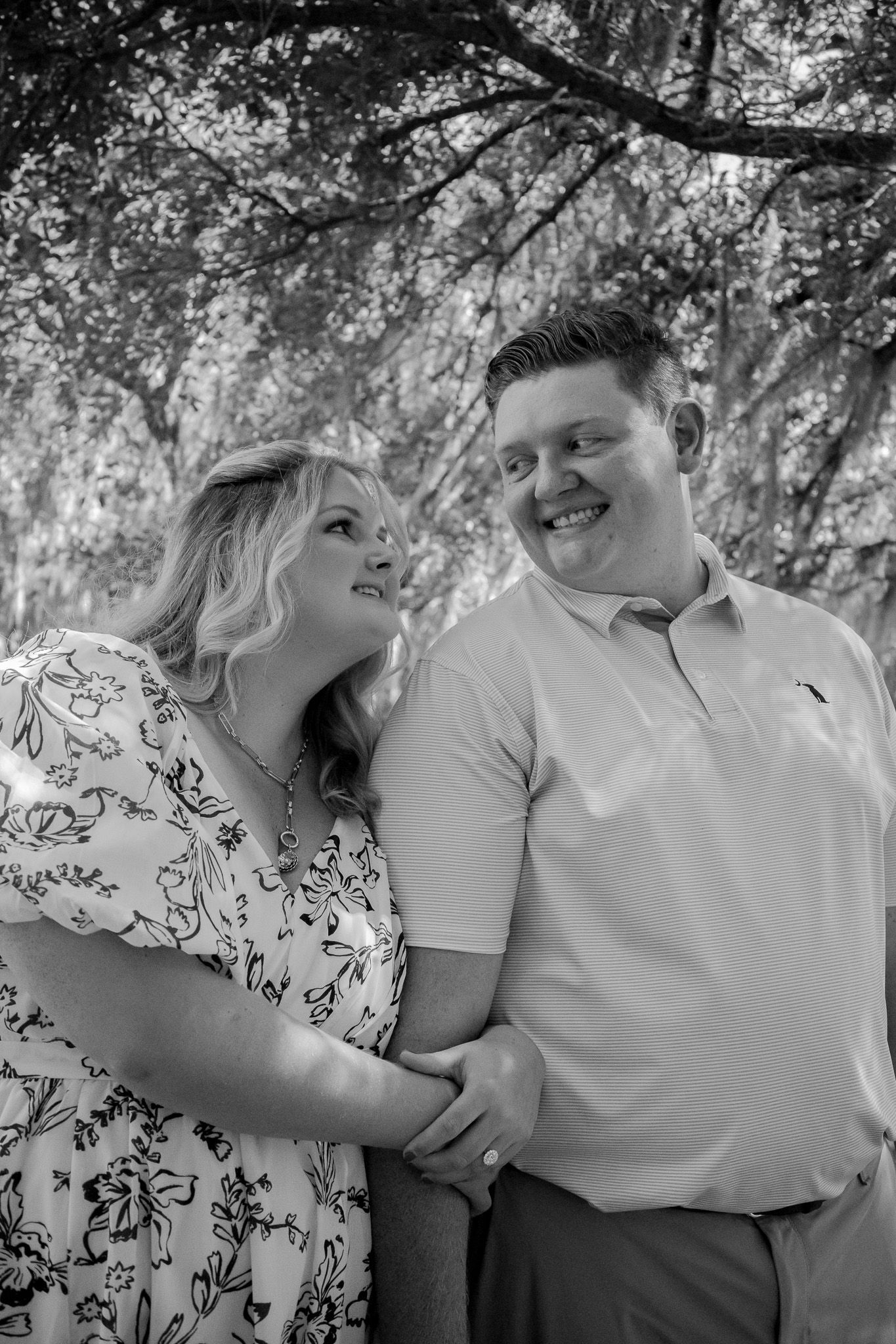 Black and white photo of a smiling couple holding hands outdoors under trees, woman in floral dress, man in polo shirt.