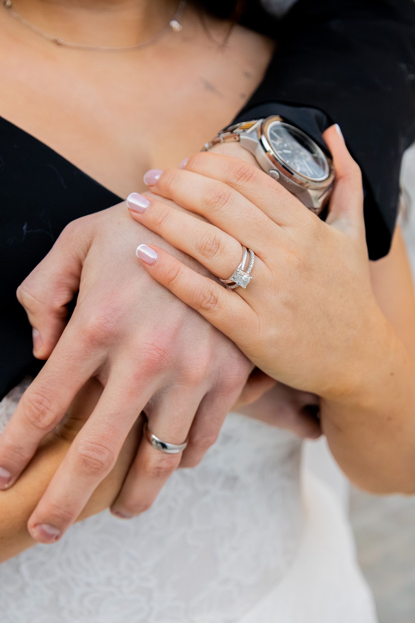 Close-up of newlyweds hands showing diamond engagement ring and wedding bands, with a watch visible.