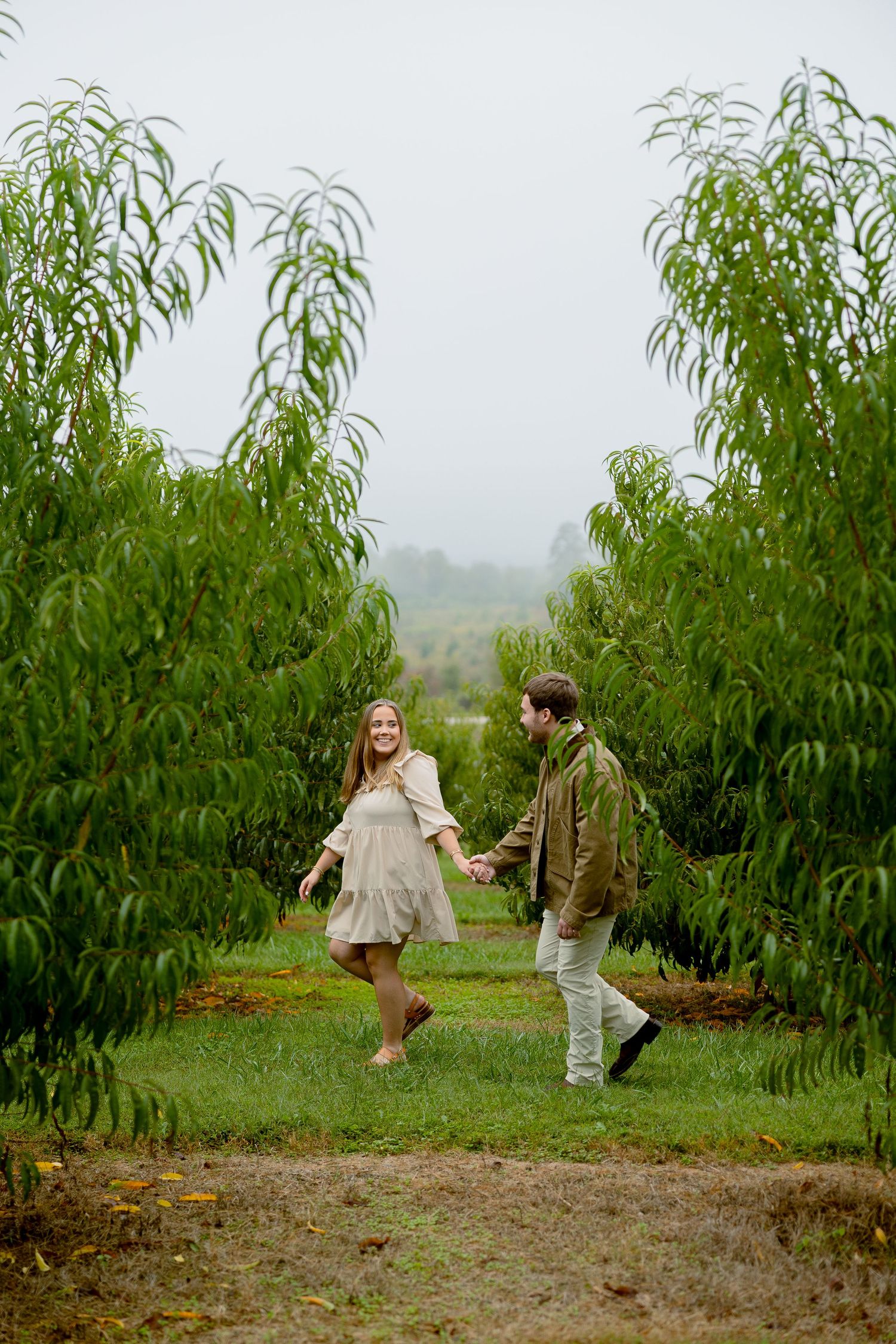 A couple holds hands and walks through a lush green orchard on a misty day, smiling joyfully.