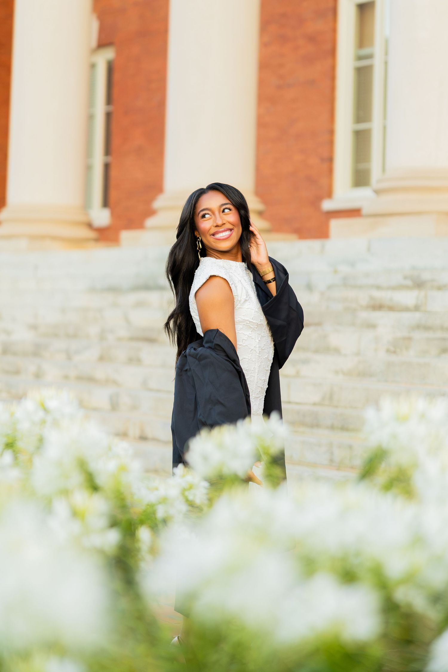 Smiling young woman in white dress and graduation gown poses by brick building steps with white flowers in foreground.