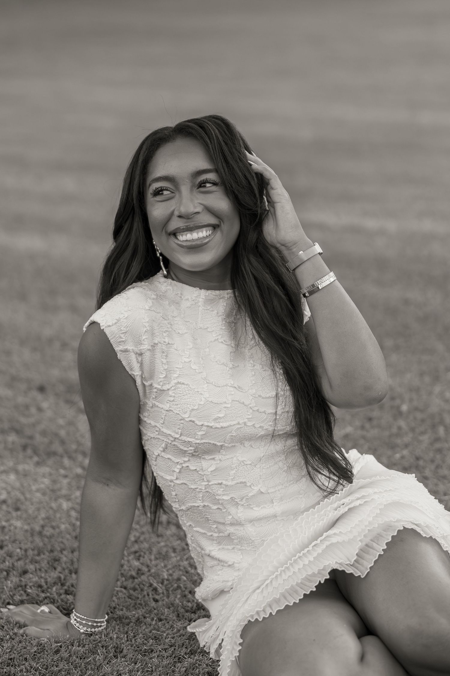 Black and white photo of a smiling young woman in a white lace dress sitting on grass, hand in hair.