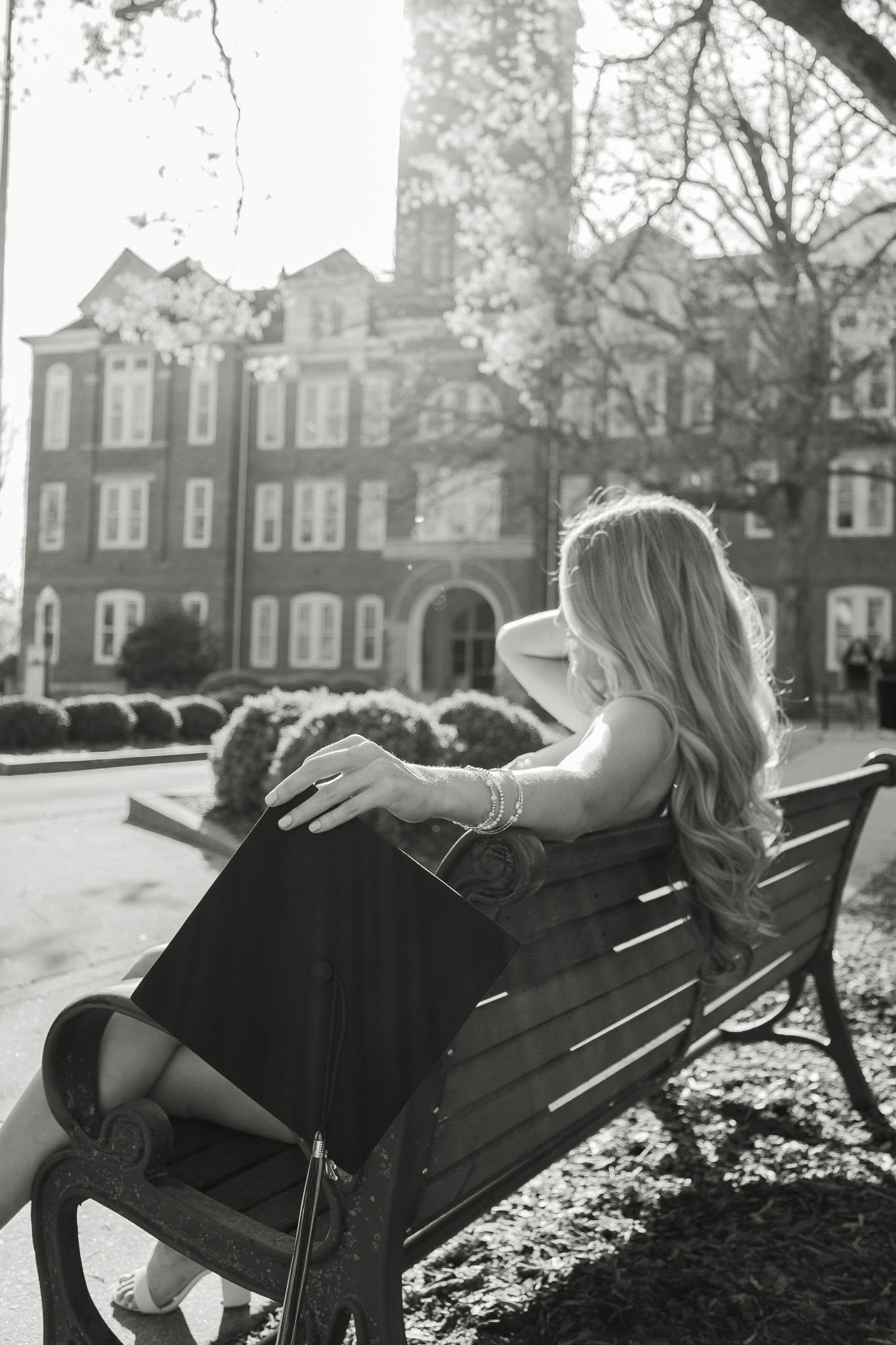 Blonde woman in elegant black dress sitting on a park bench in front of a classic brick university building.