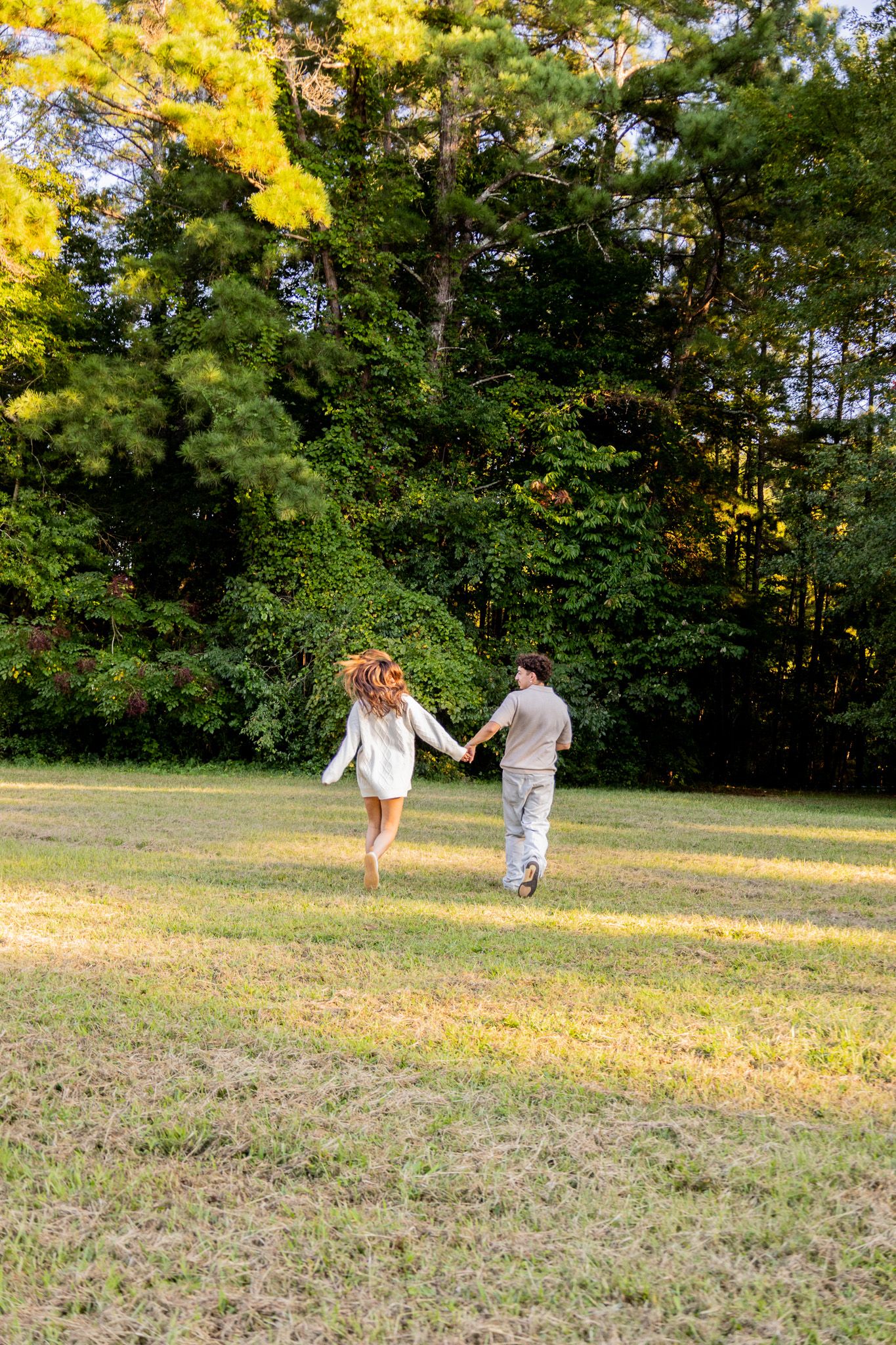 A couple holds hands and runs through a sunlit green field surrounded by lush trees.