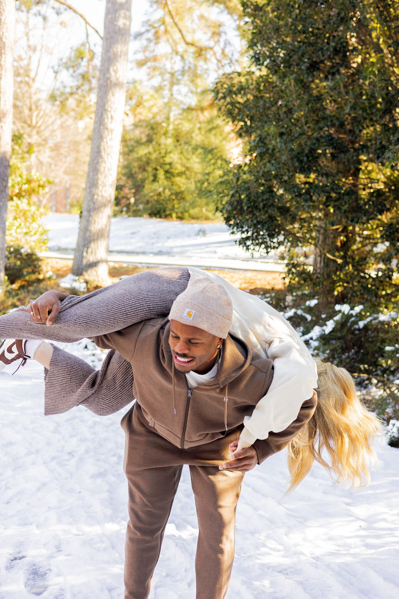 A smiling man carries a laughing blonde woman over his shoulder in a snowy outdoor setting on a sunny winter day.