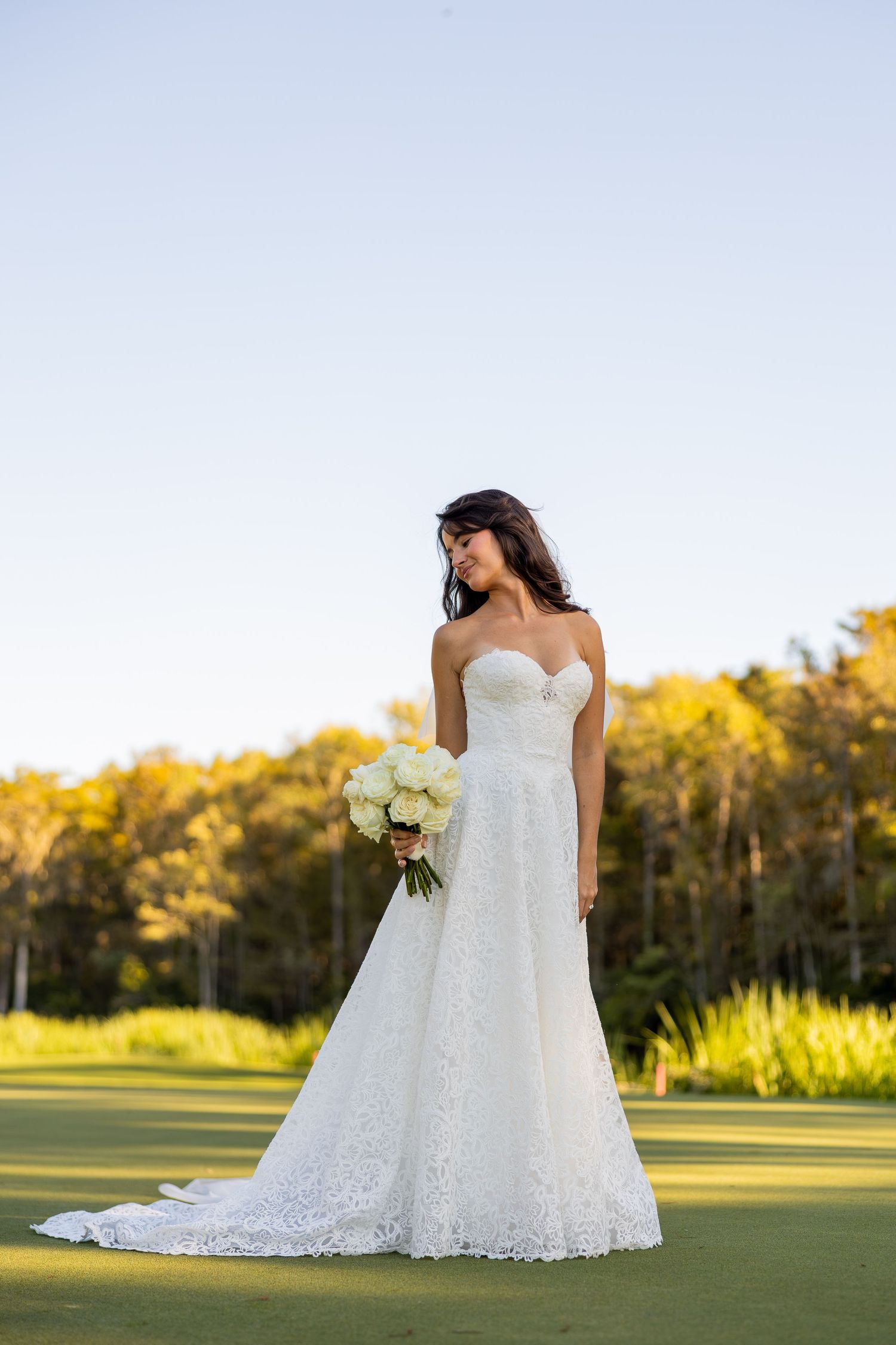 Elegant bride in strapless lace A-line gown holds white bouquet on green lawn at golden hour.