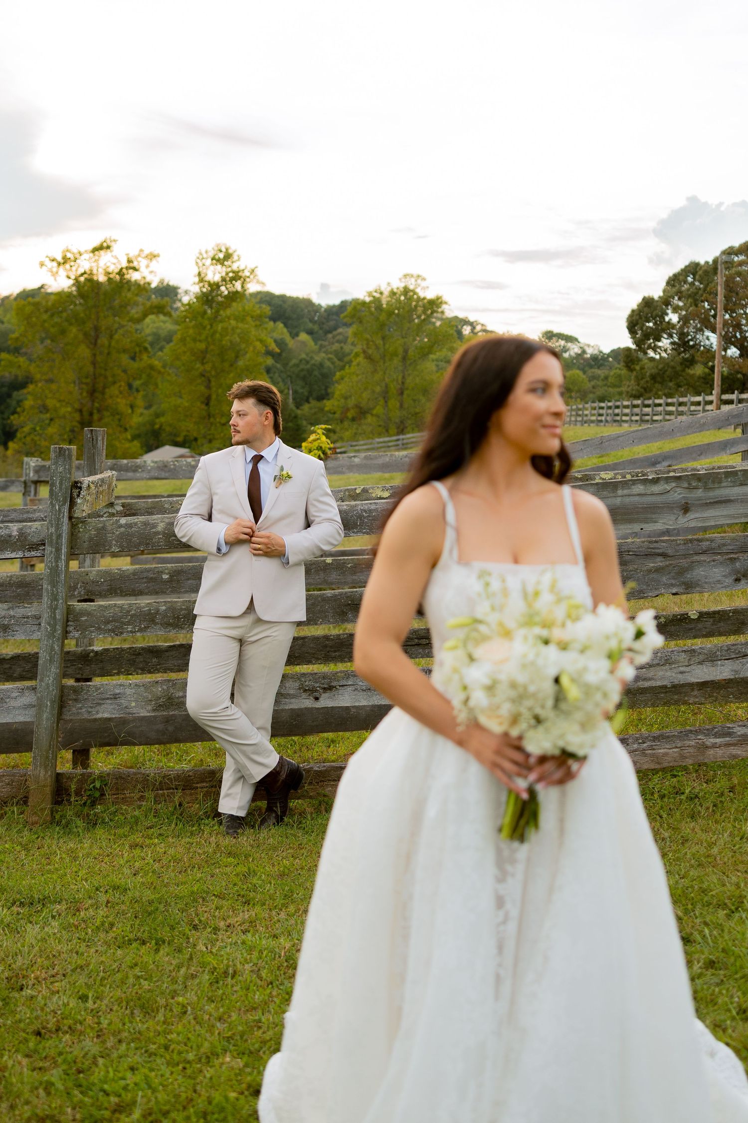 Bride in white gown holding bouquet with groom in tan suit leaning on rustic fence at outdoor wedding.
