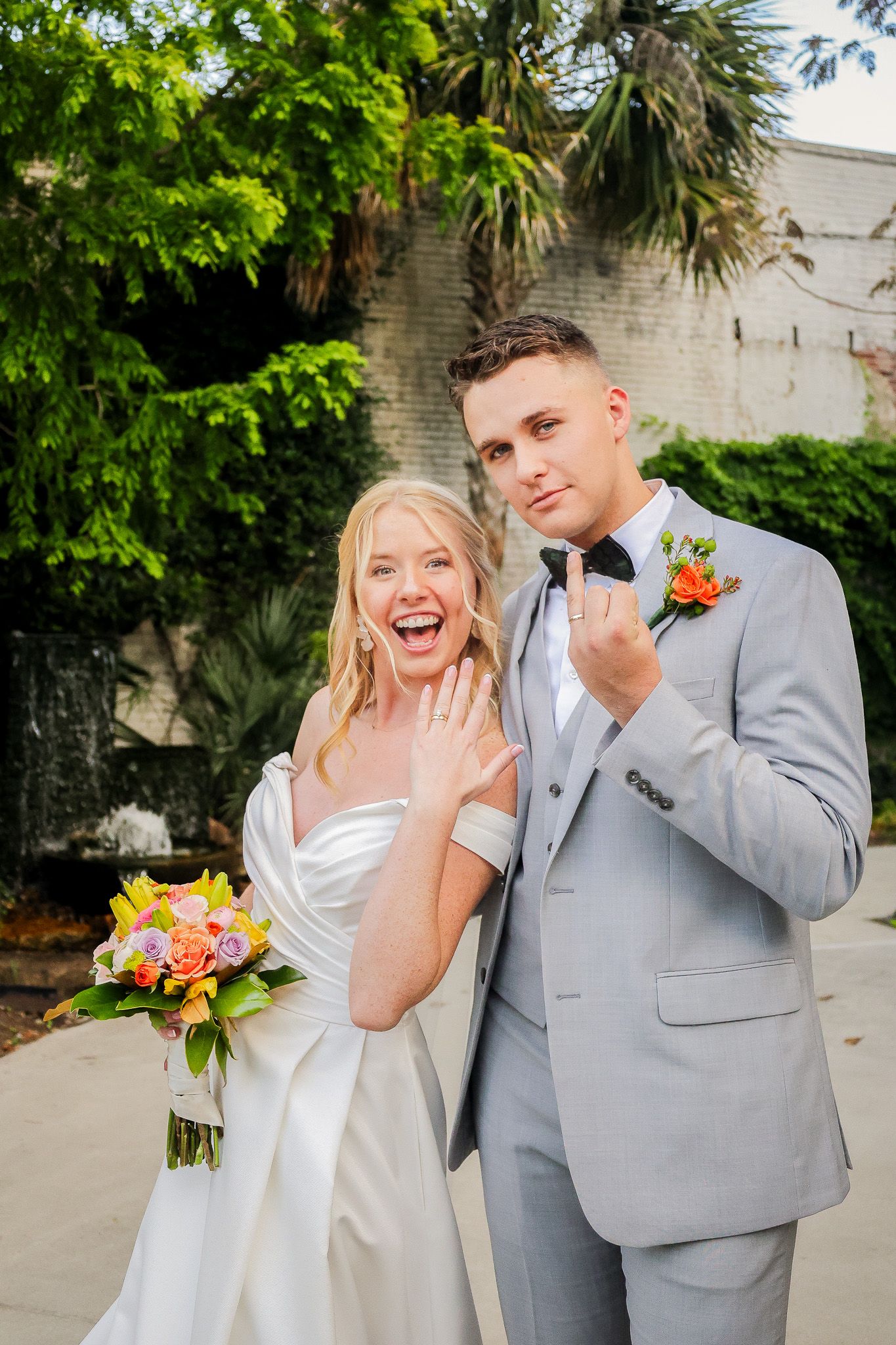 Joyful bride shows off wedding ring beside groom in gray suit holding colorful bouquet outdoors.