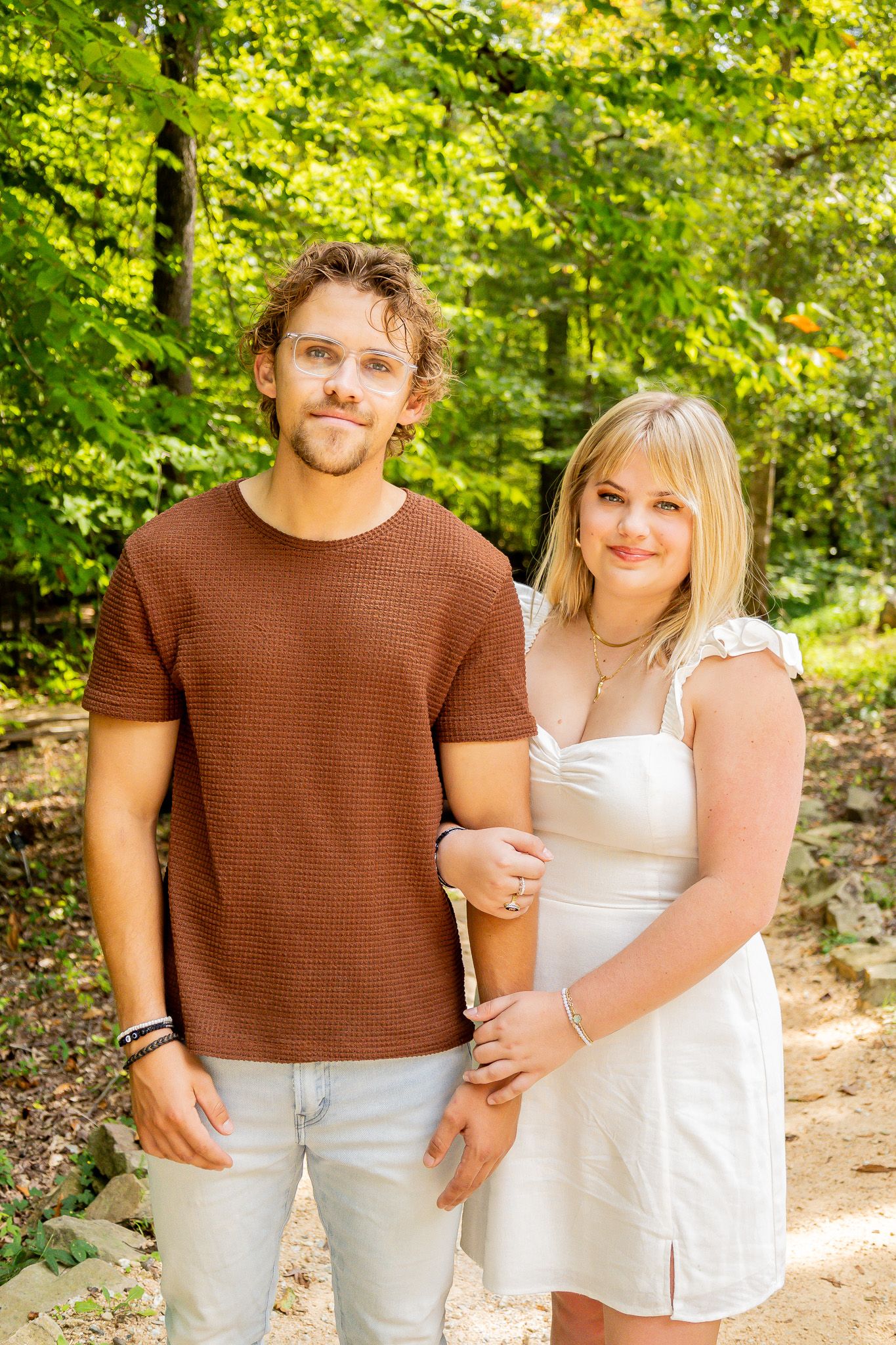 A young couple poses outdoors in a lush green forest, man in brown shirt and woman in white dress.