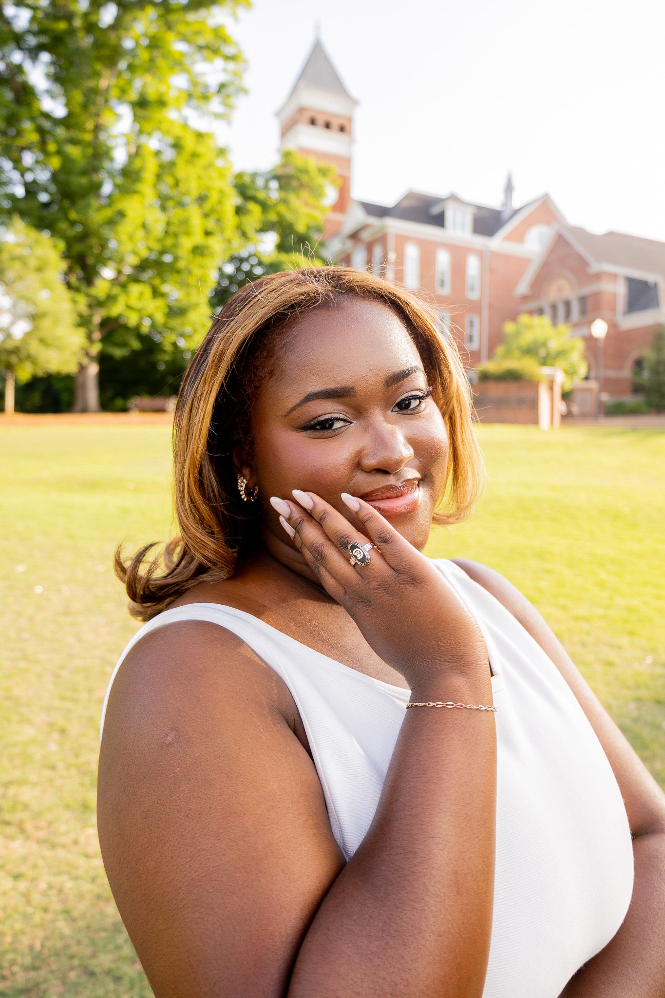 Young Black woman in white dress smiles gracefully with hand on chin near brick university building.