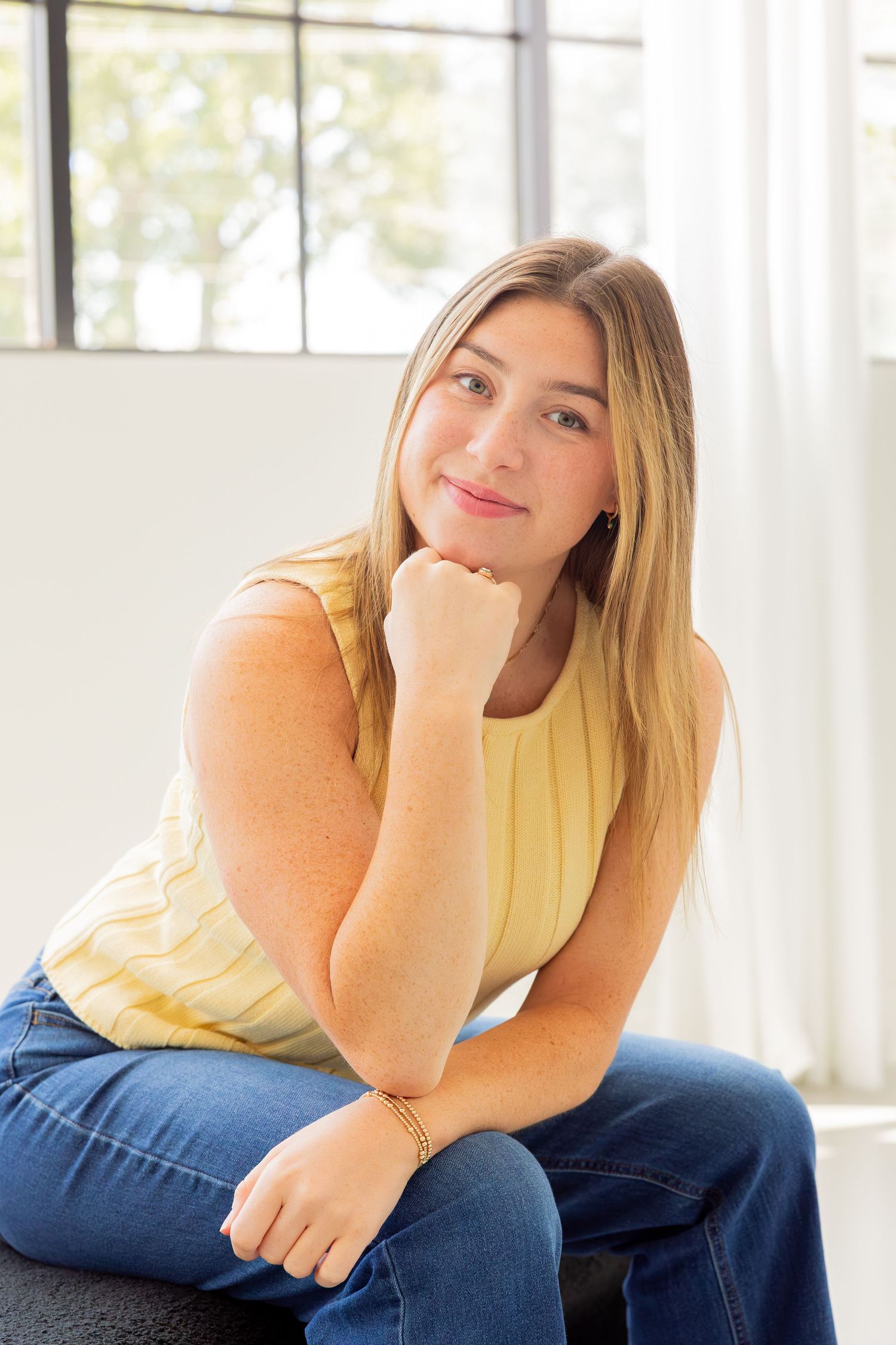 Young woman with blonde hair in yellow top and jeans sits smiling with chin resting on fist.