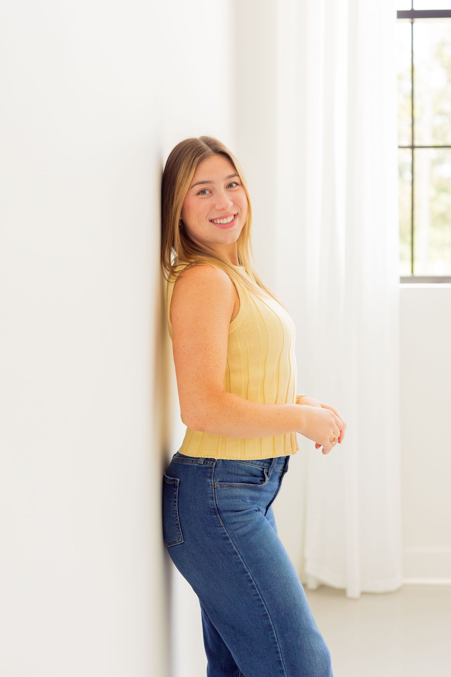 Smiling young woman in yellow tank top and blue jeans leaning against a white wall near a bright window.