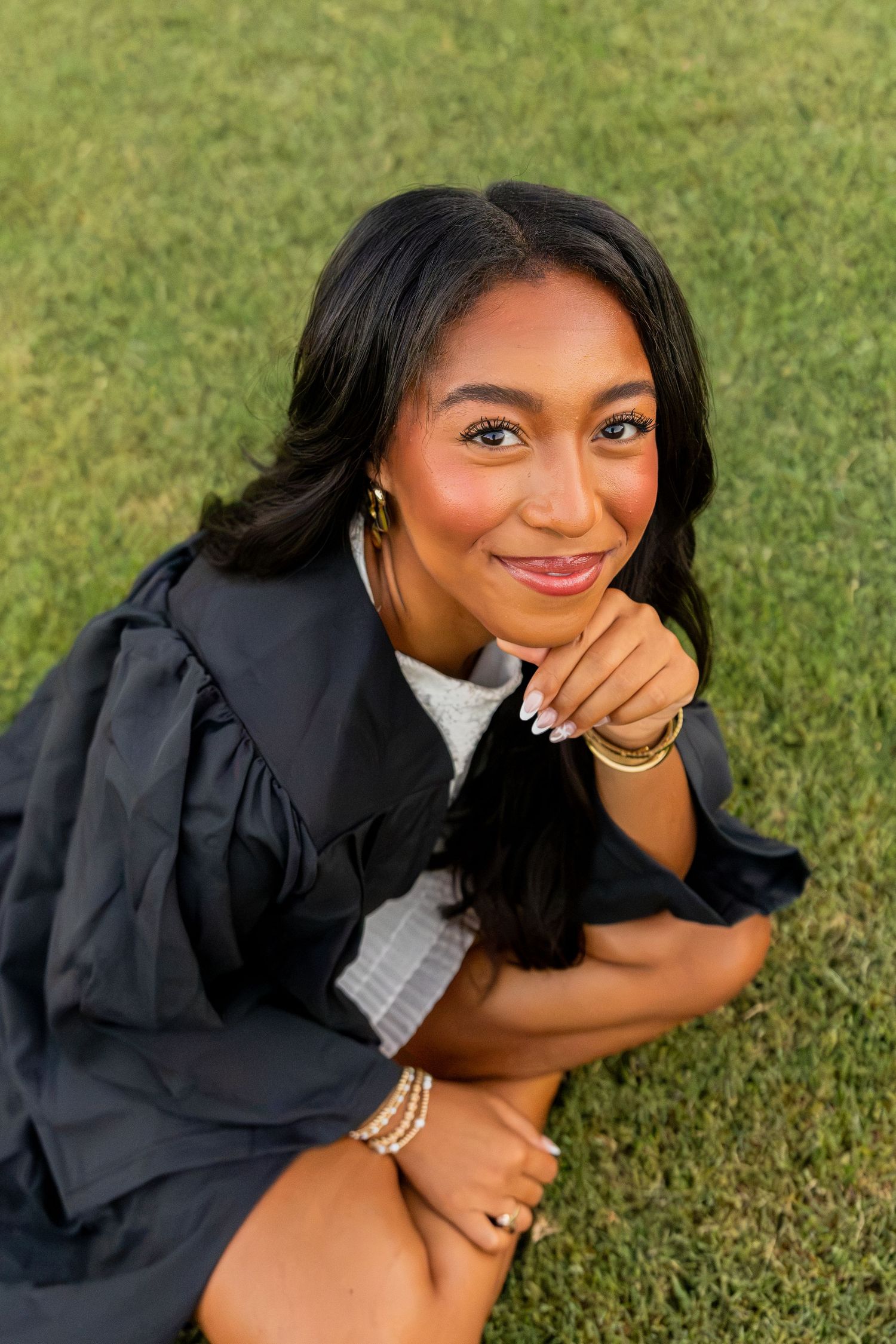 Close-up portrait of smiling graduate in black gown with gold jewelry sitting on green grass.