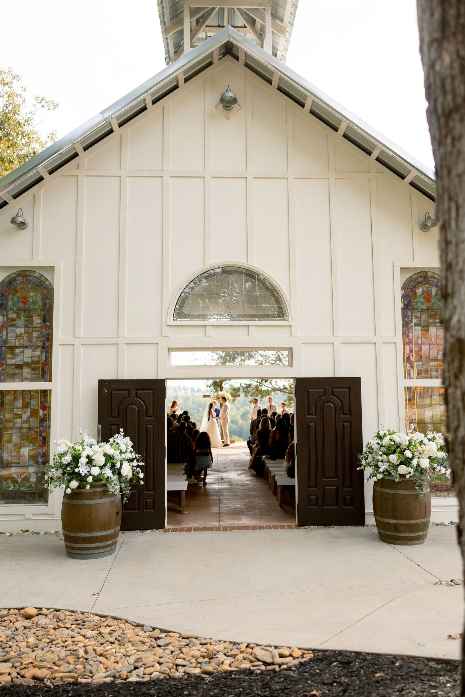 White chapel wedding venue with open doors, barrel planters with white flowers, and ceremony visible inside.