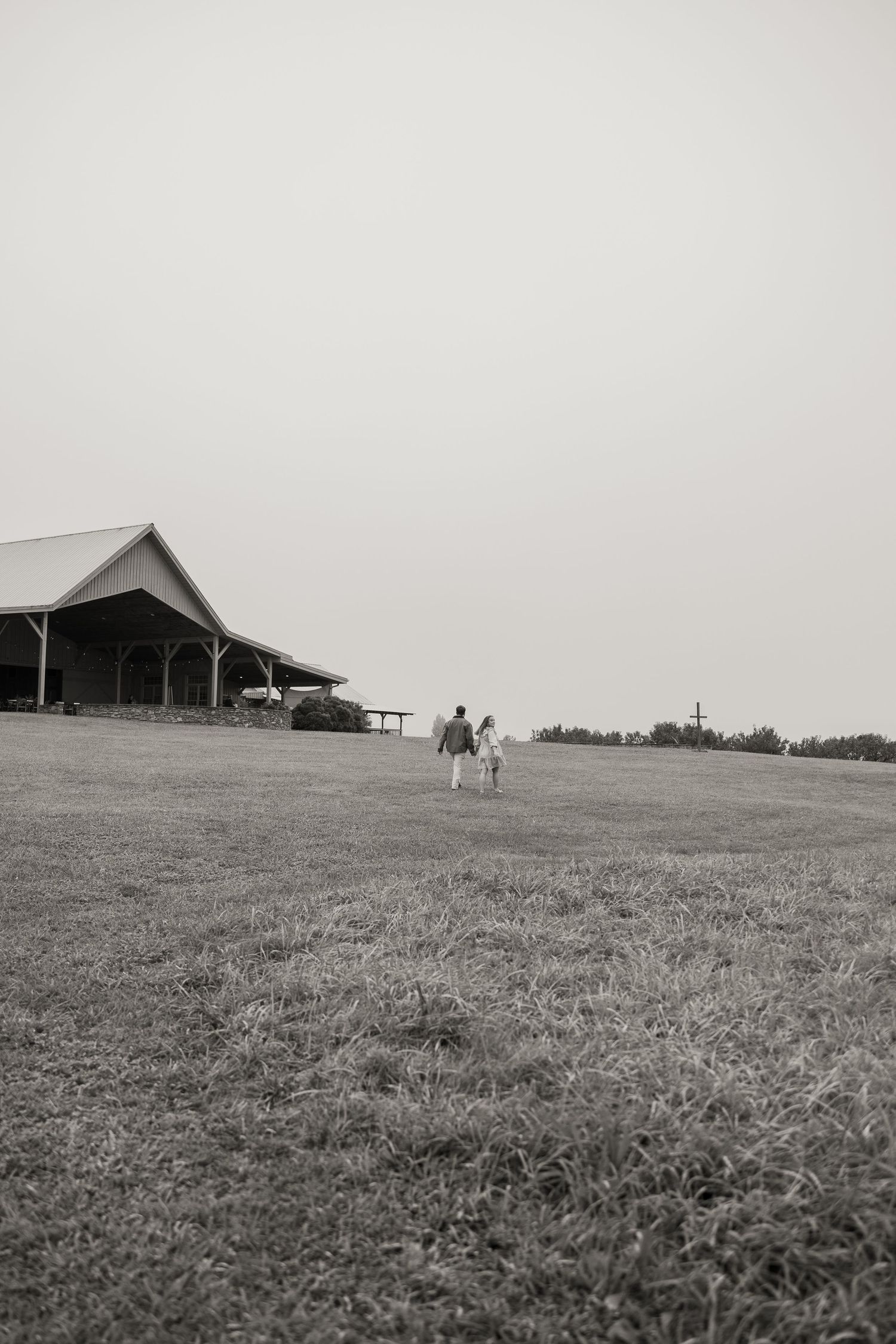 Black and white photo of a couple holding hands walking toward a large barn venue across an open grassy field.