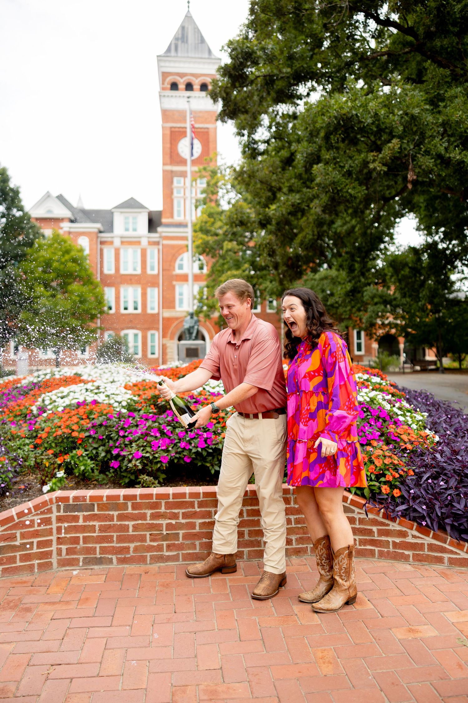 Couple celebrating with champagne by colorful flower garden near brick clock tower building on college campus.