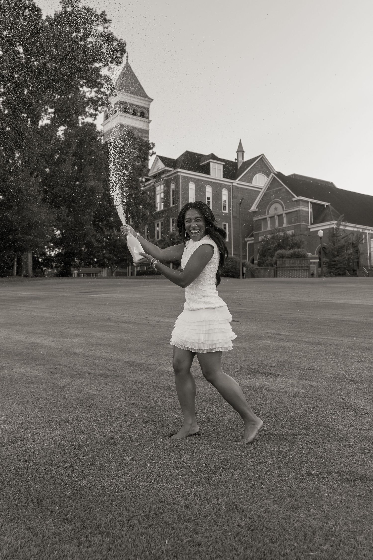 Young woman in white dress dancing barefoot on grass in front of historic brick building with tower, black and white photo.