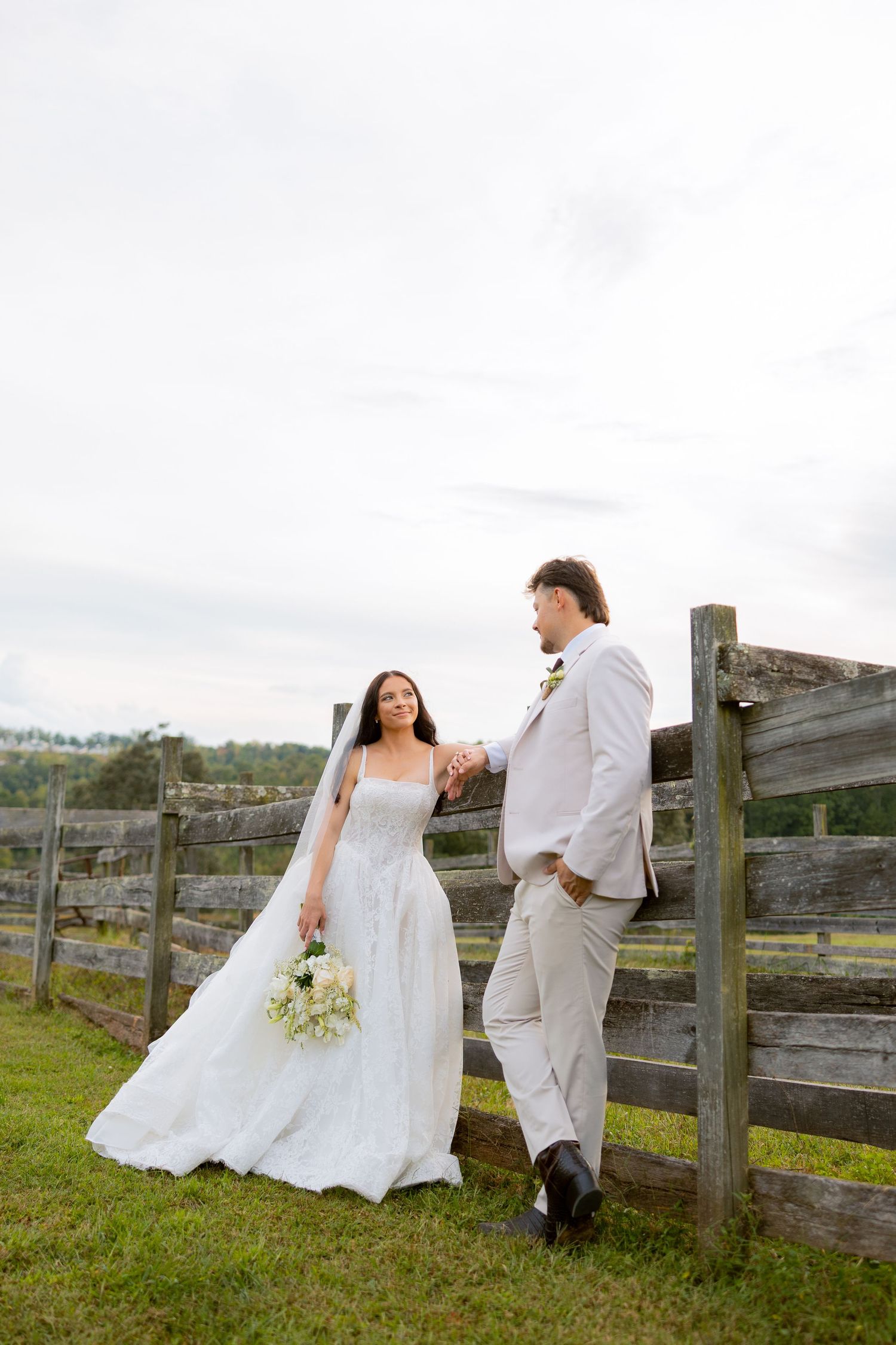 Bride in white gown and groom in tan suit hold hands by rustic wooden fence on green farm landscape.