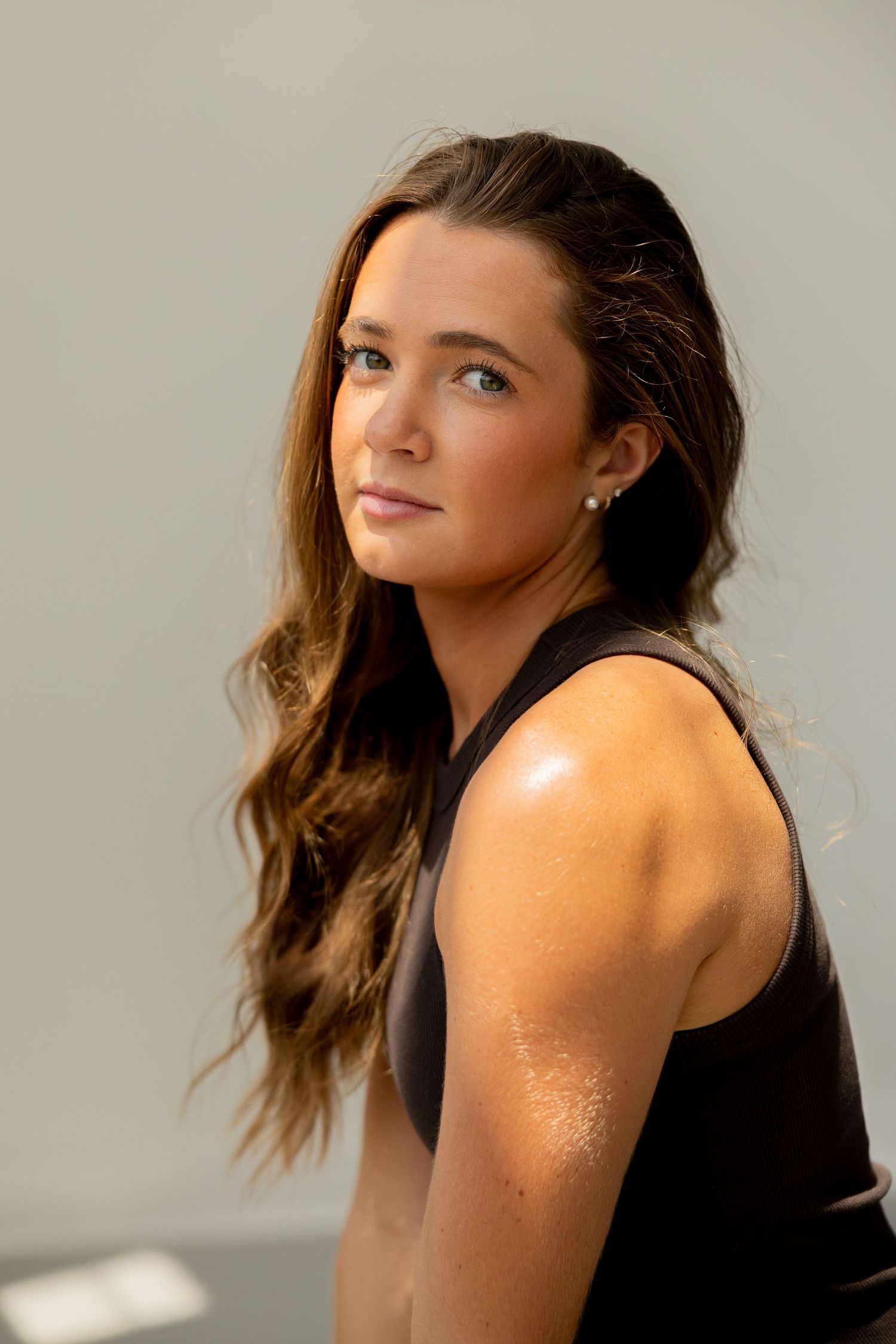 Young woman with long brown hair wearing a black sleeveless top, posing confidently against a gray background.
