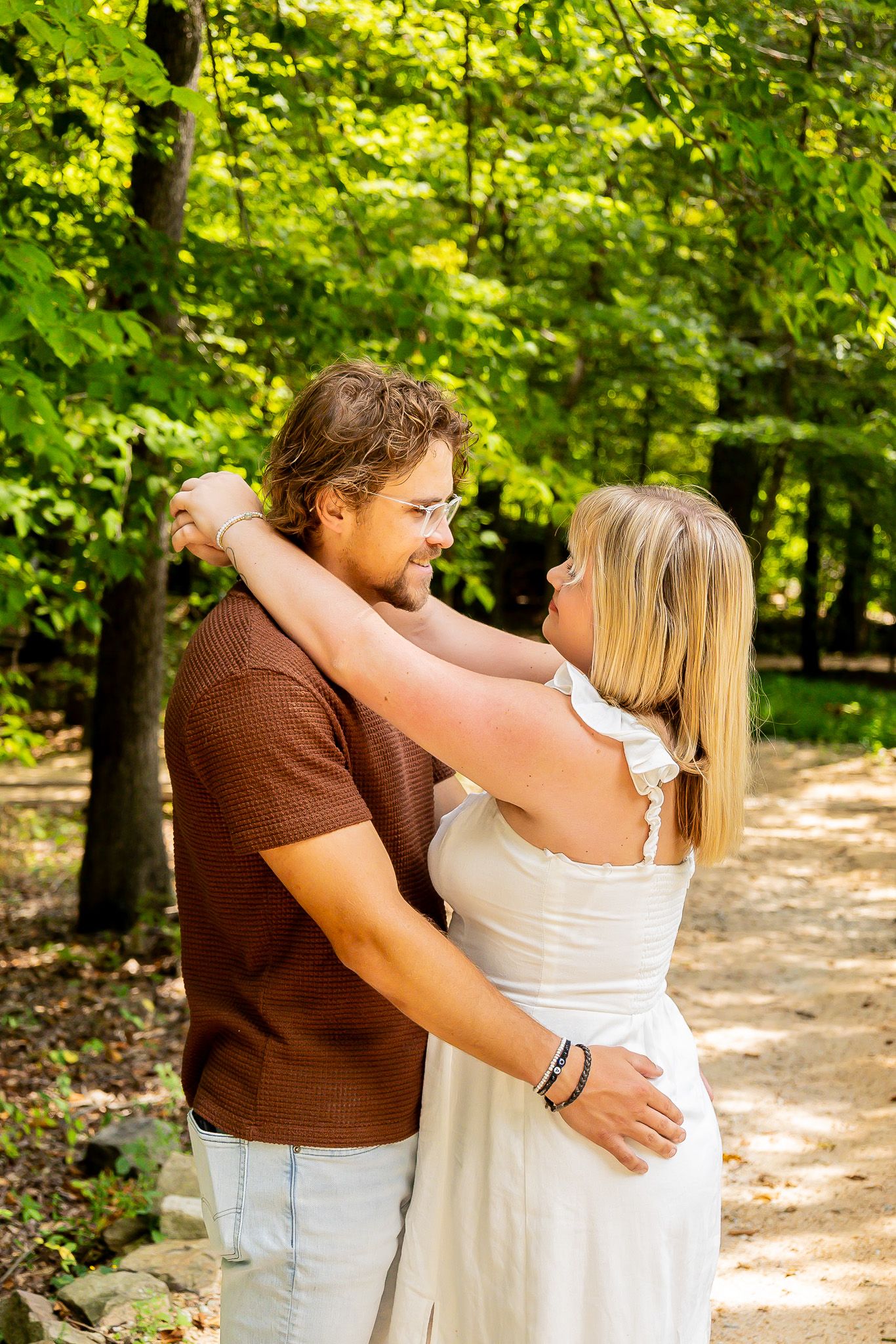 Couple embracing on a sunny forest path, woman in white dress with arms around man in brown shirt.