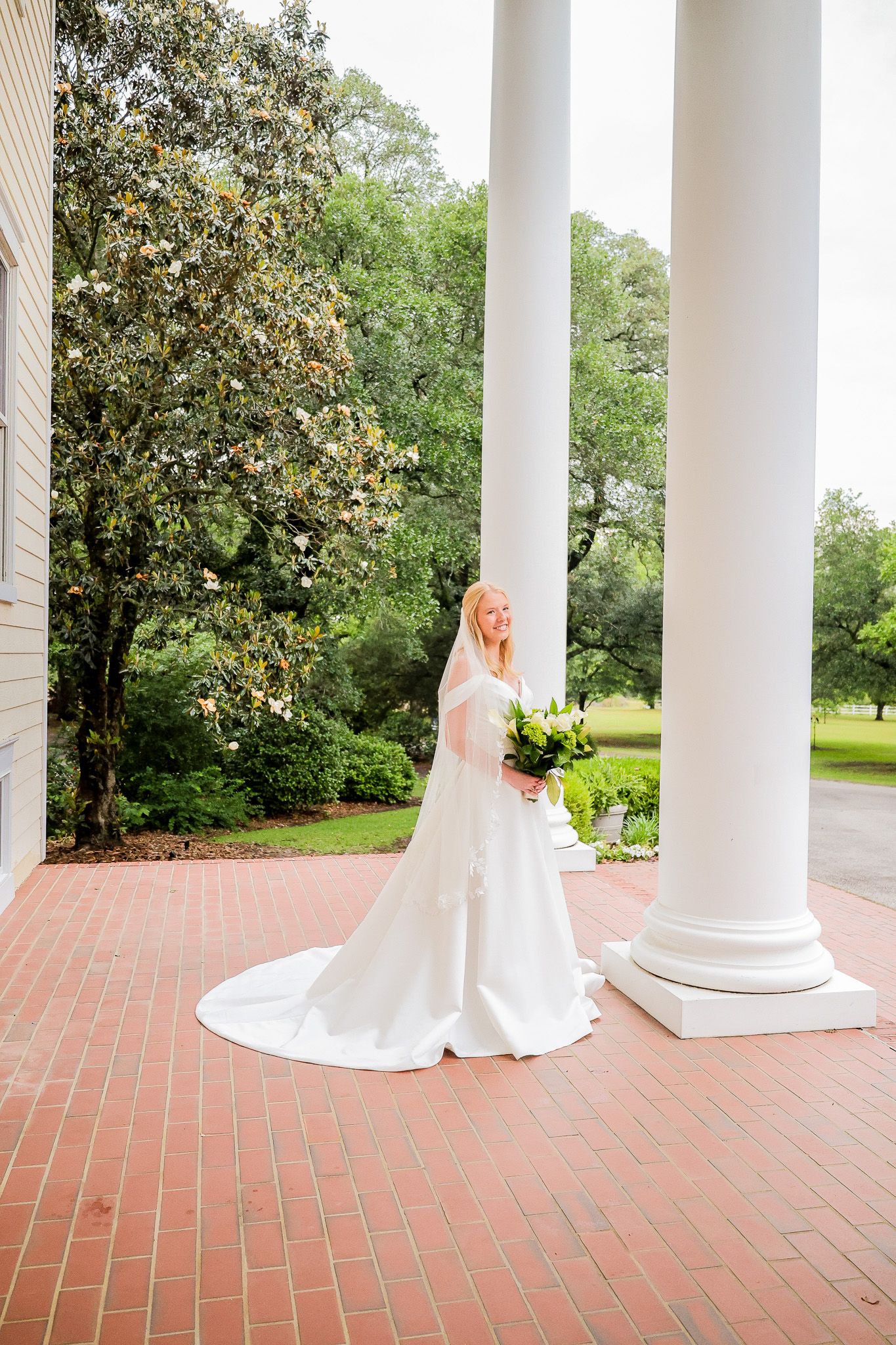 Bride in white gown with train holds bouquet beside tall white columns on brick porch with lush green trees.
