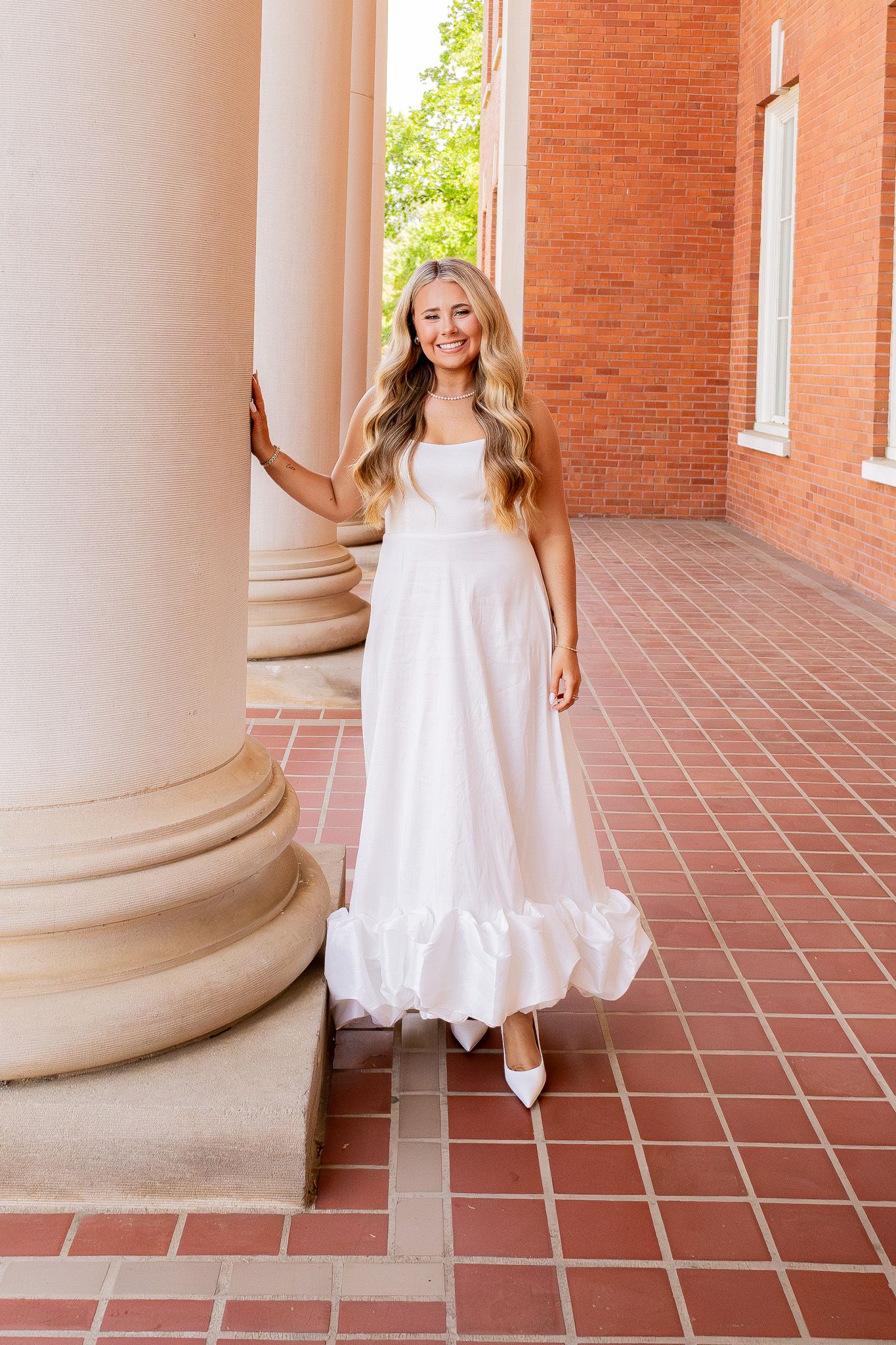 Smiling blonde woman in white ruffled maxi dress and heels posing by columns of a brick building.