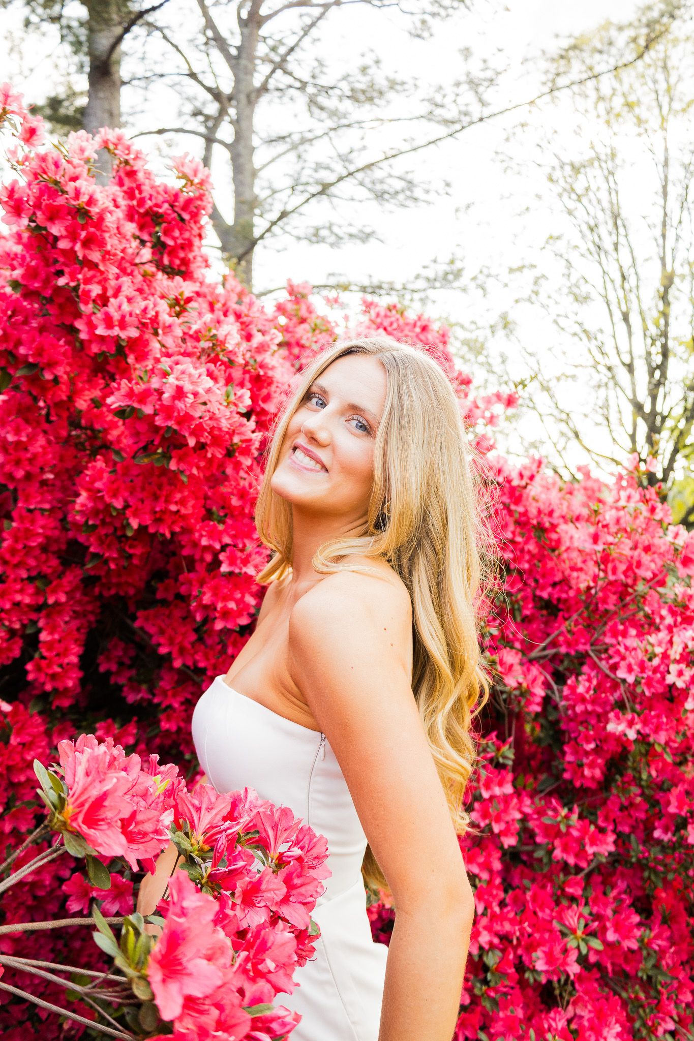 Beautiful blonde woman smiling among vibrant pink azalea blooms in a white strapless dress outdoors.