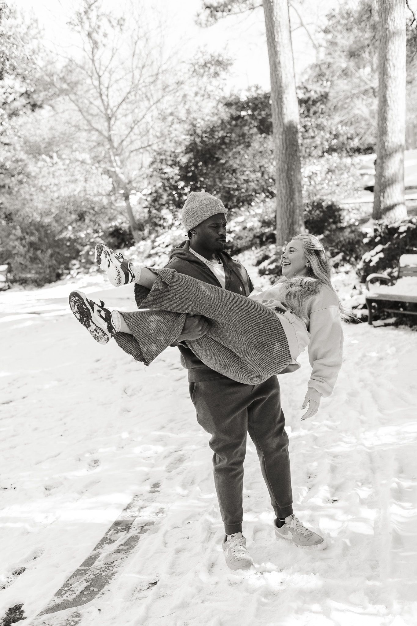 Man carrying woman in his arms laughing together in a snowy winter wonderland setting, black and white photo.