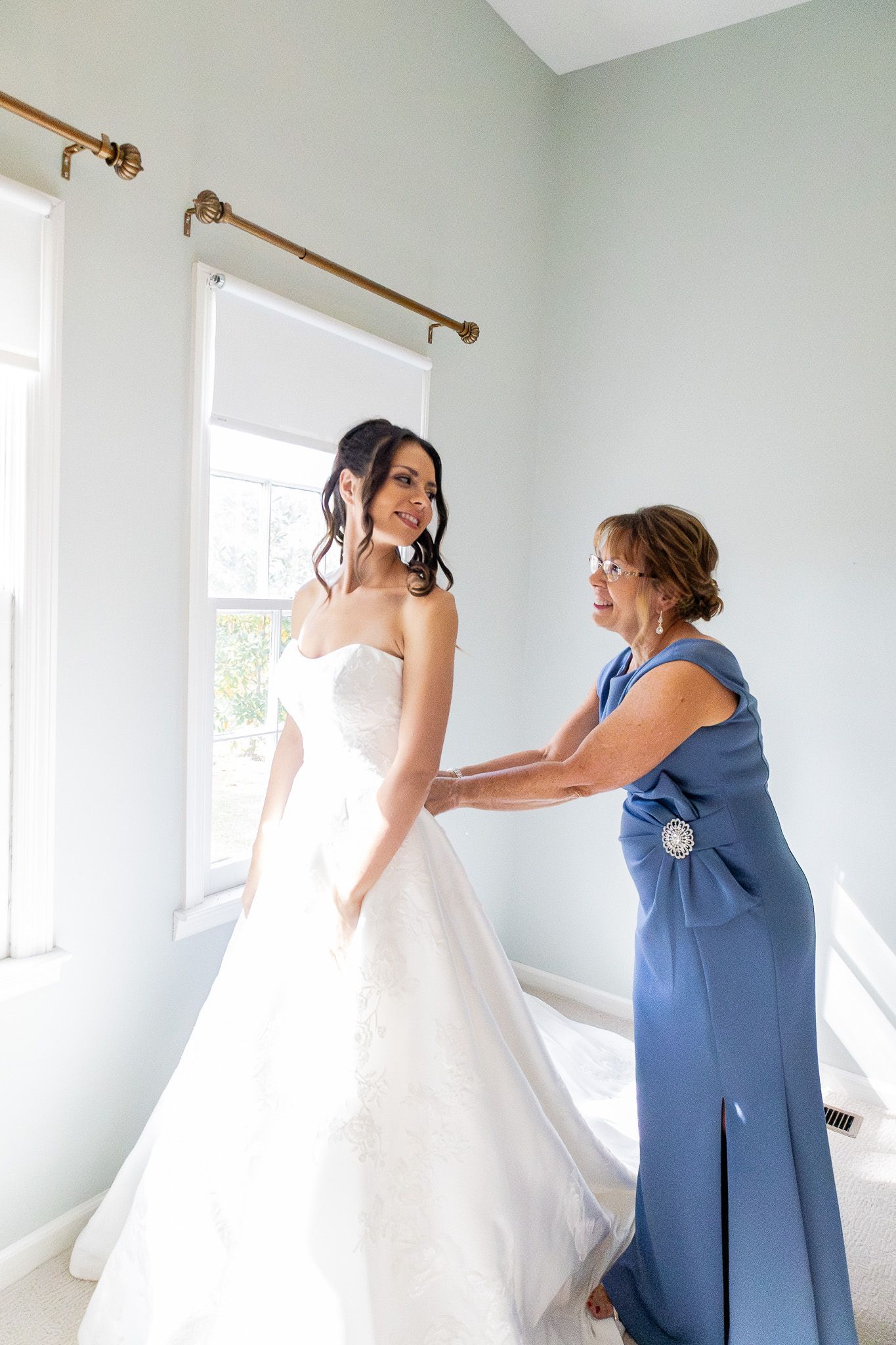 Bride in white gown smiles as woman in blue dress helps fasten her wedding dress in a bright, elegant room.