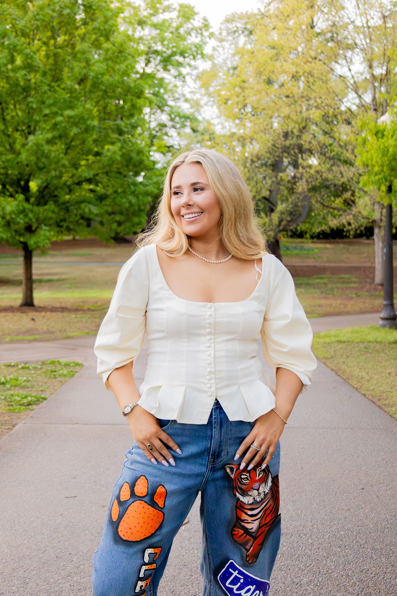 Smiling blonde woman in white top and Clemson Tigers painted jeans posing outdoors in a park.