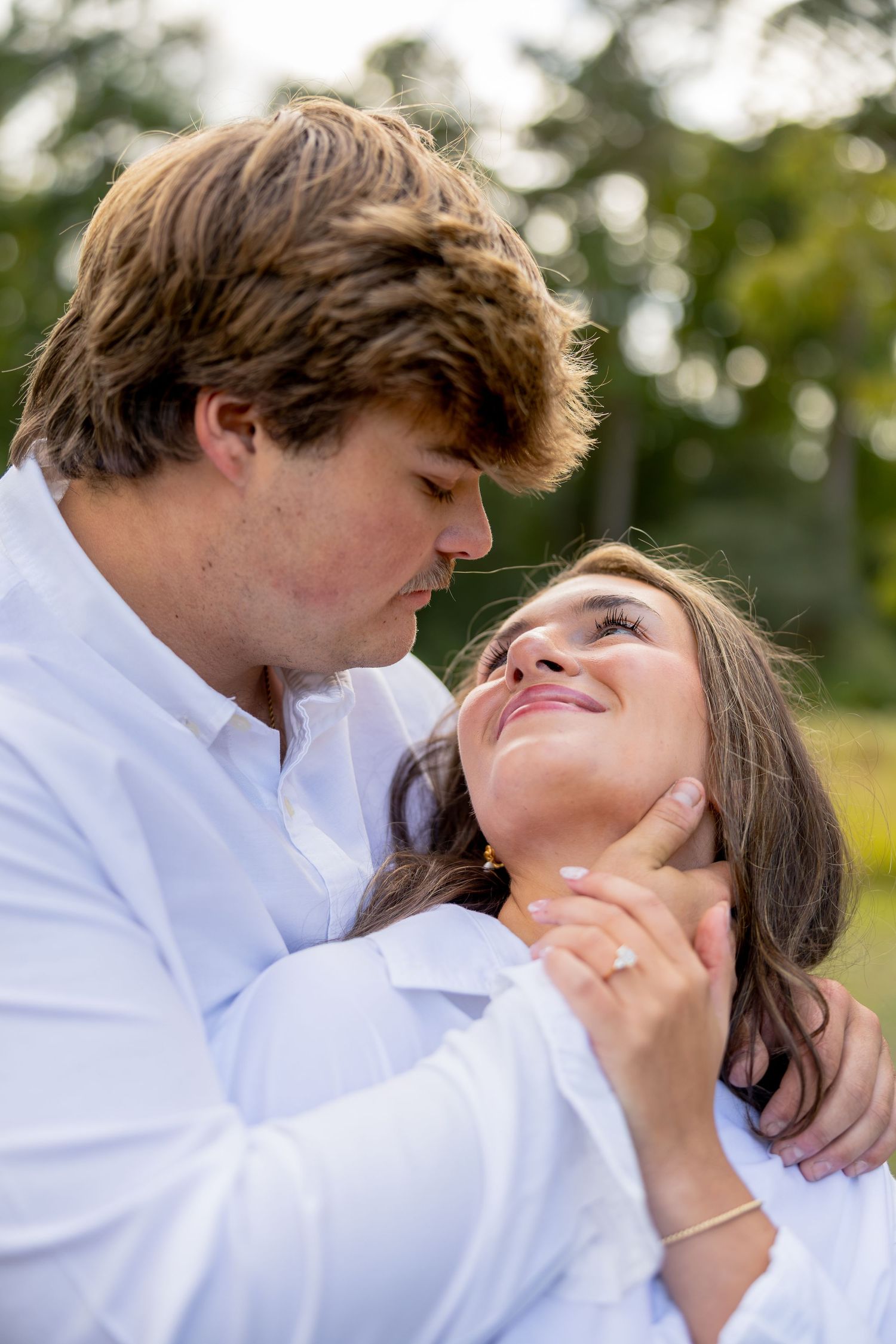 Couple in white shirts embracing outdoors as the woman smiles up at the man during a romantic engagement photo session.