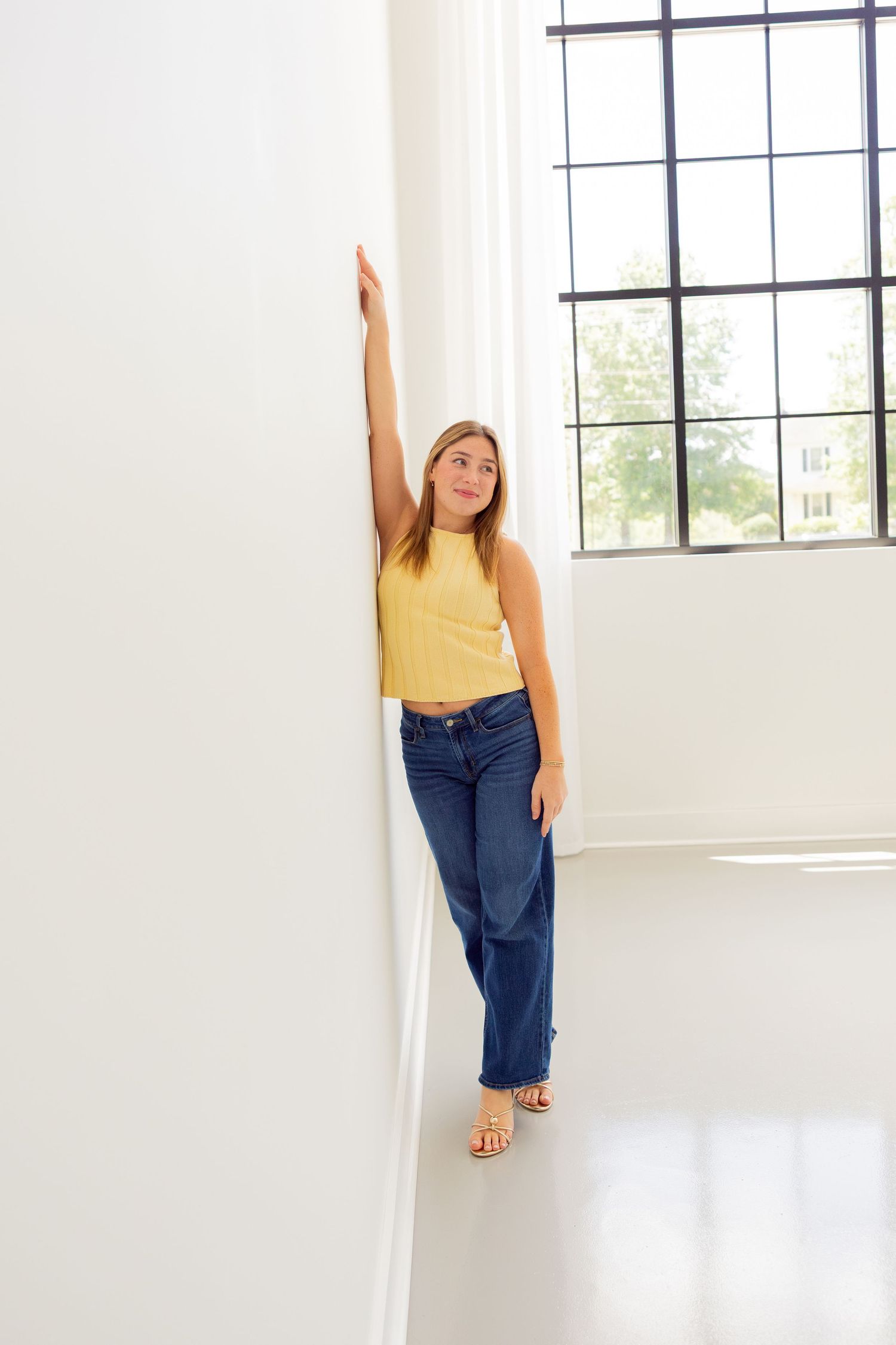 Young woman in yellow crop top and wide-leg jeans leaning against white wall near large window.