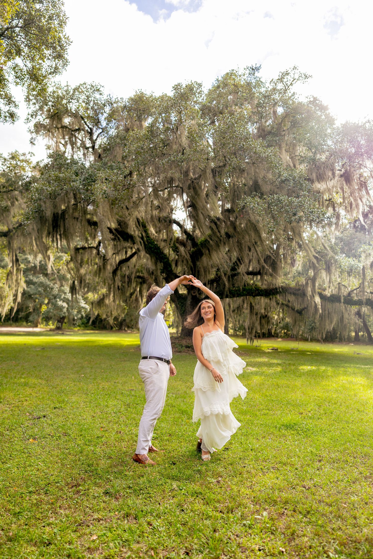 Couple in white twirling and dancing on a sunny green lawn beneath a large live oak tree draped with Spanish moss.