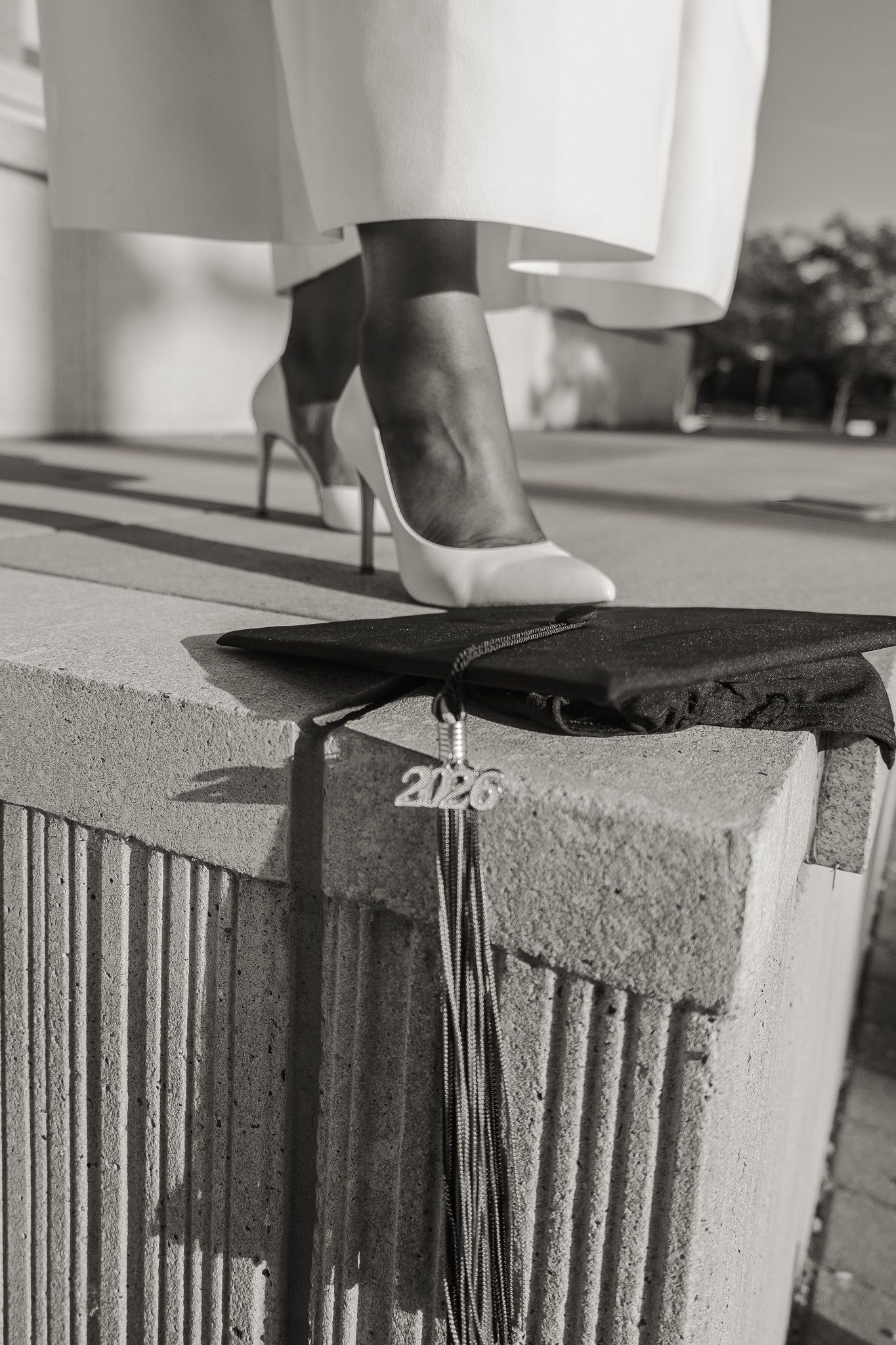 Black and white photo of graduation cap with 2023 tassel on concrete ledge beside woman in heels.