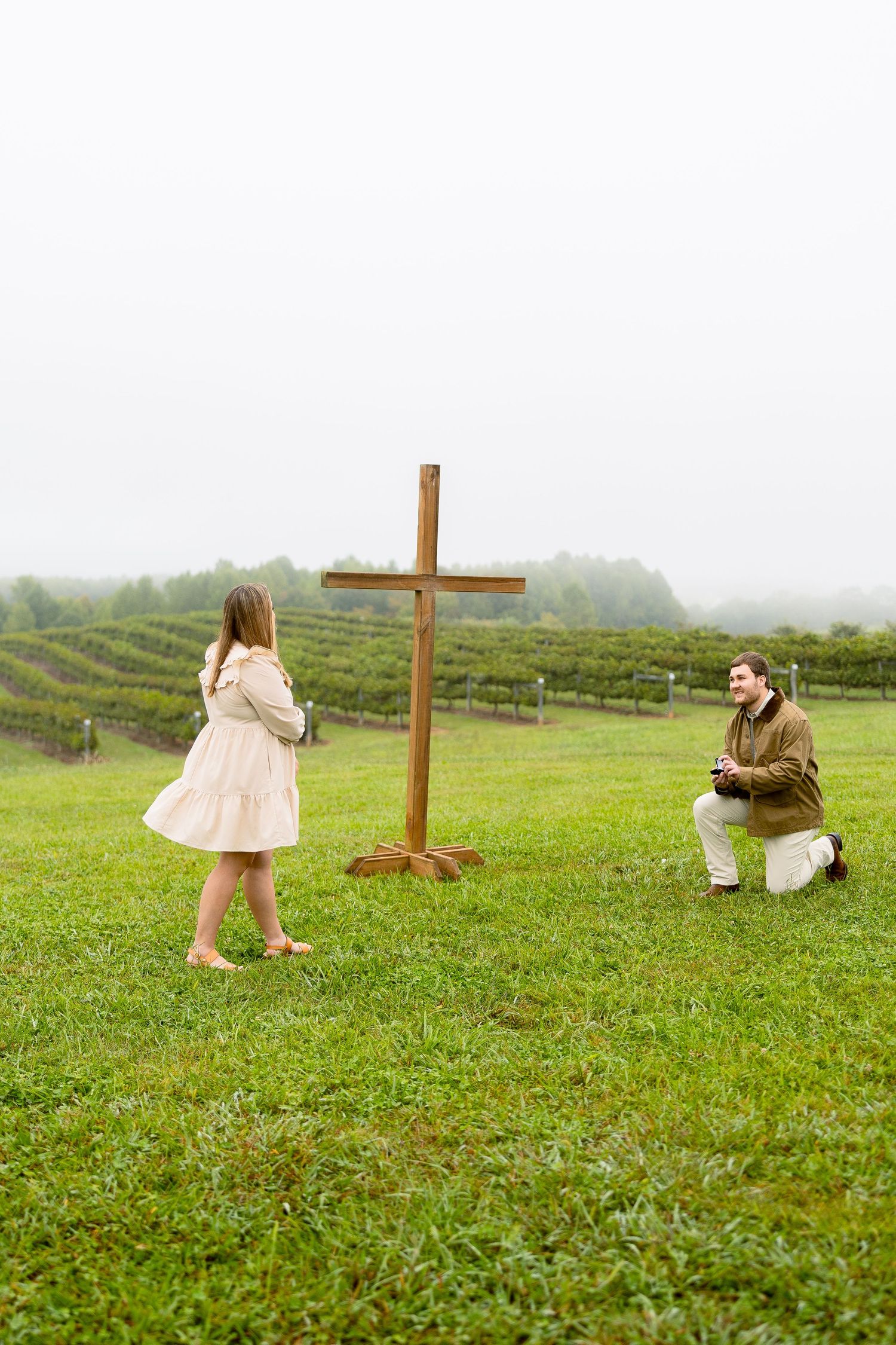 Man kneeling in proposal before woman near wooden cross in lush green vineyard on a misty day.