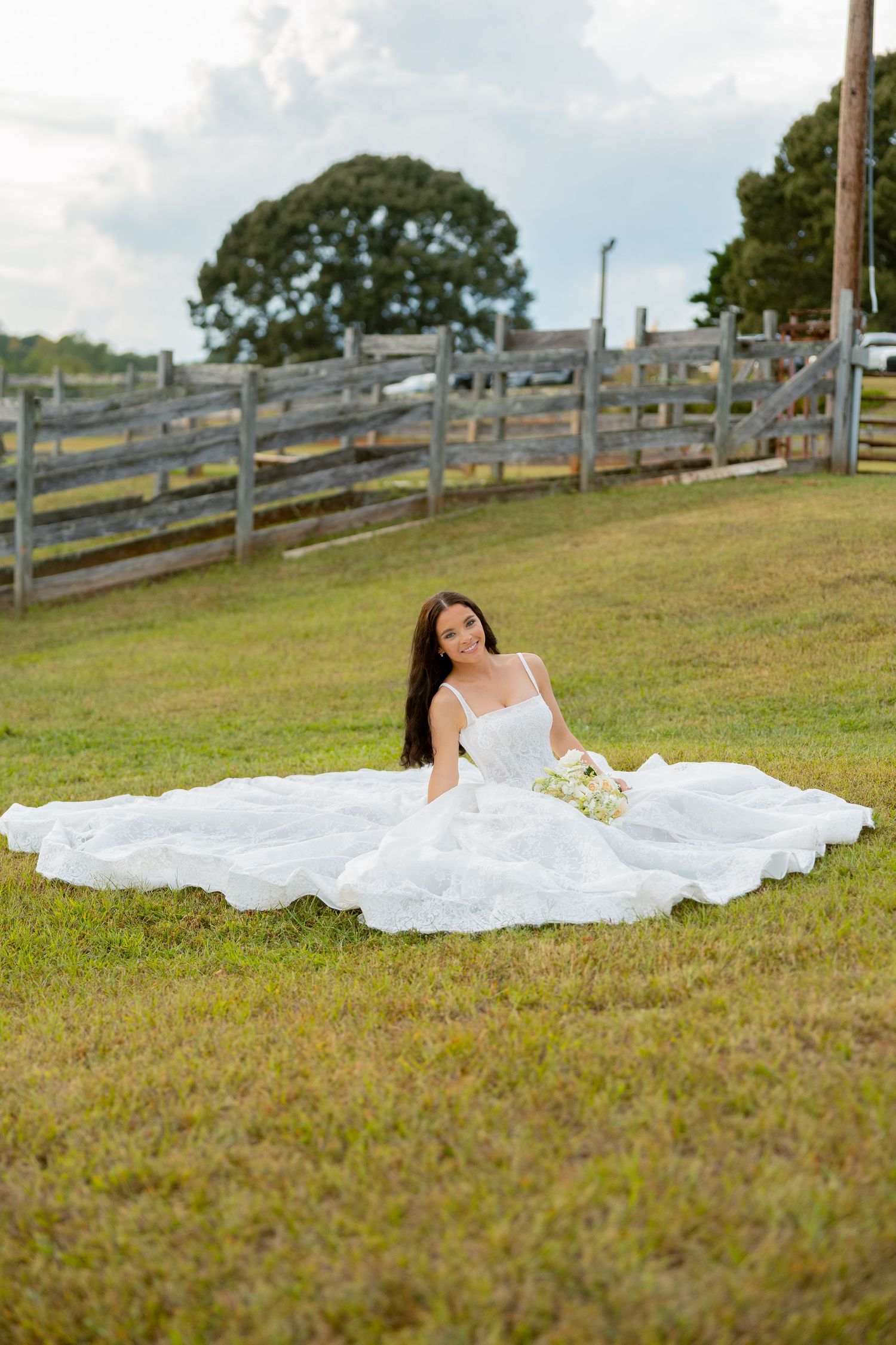 Bride in a flowing white gown sits gracefully on green grass near a wooden fence, holding a bouquet.