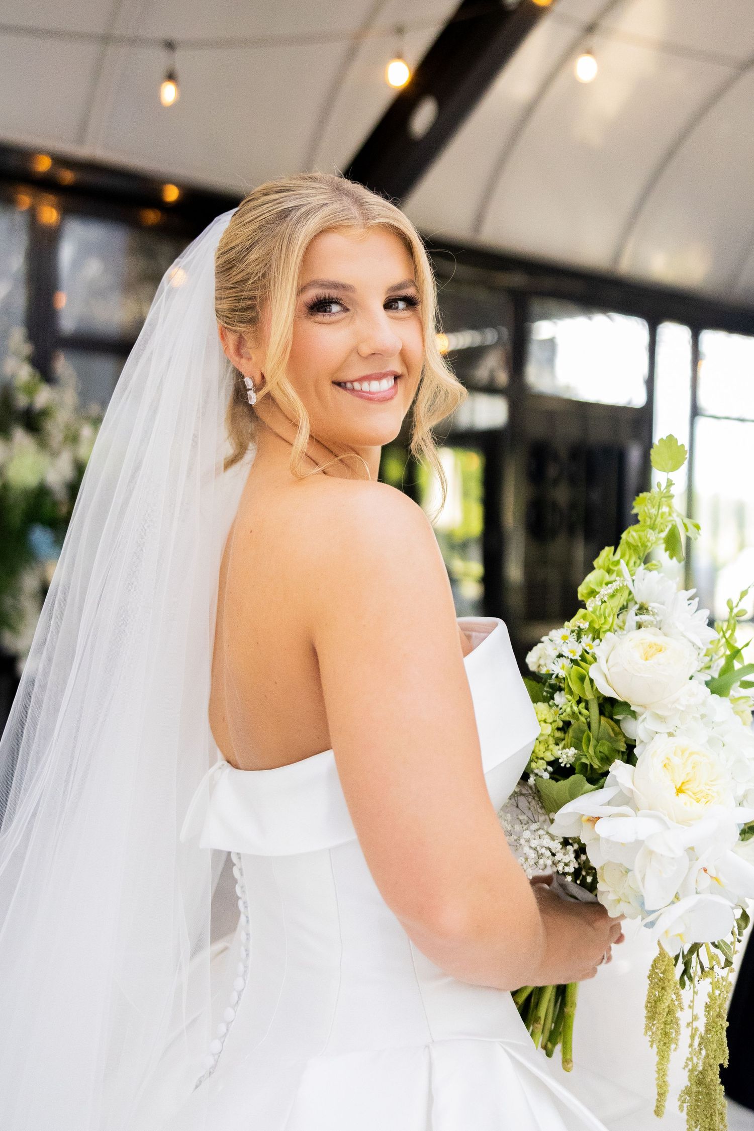 Smiling bride in strapless white gown and veil holds white floral bouquet, posing elegantly at a decorated wedding venue.