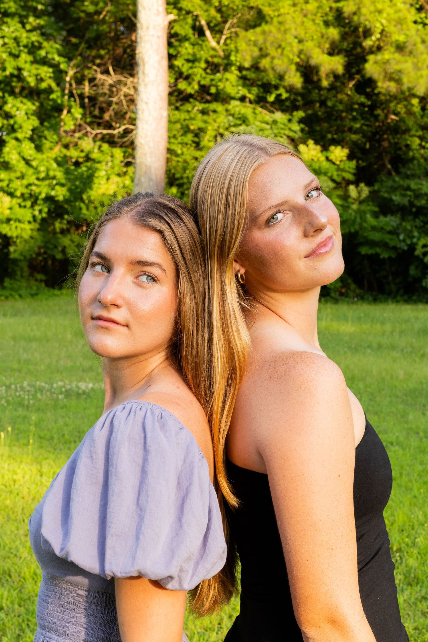 Two young women with long hair pose back-to-back outdoors in a lush green park during golden hour.
