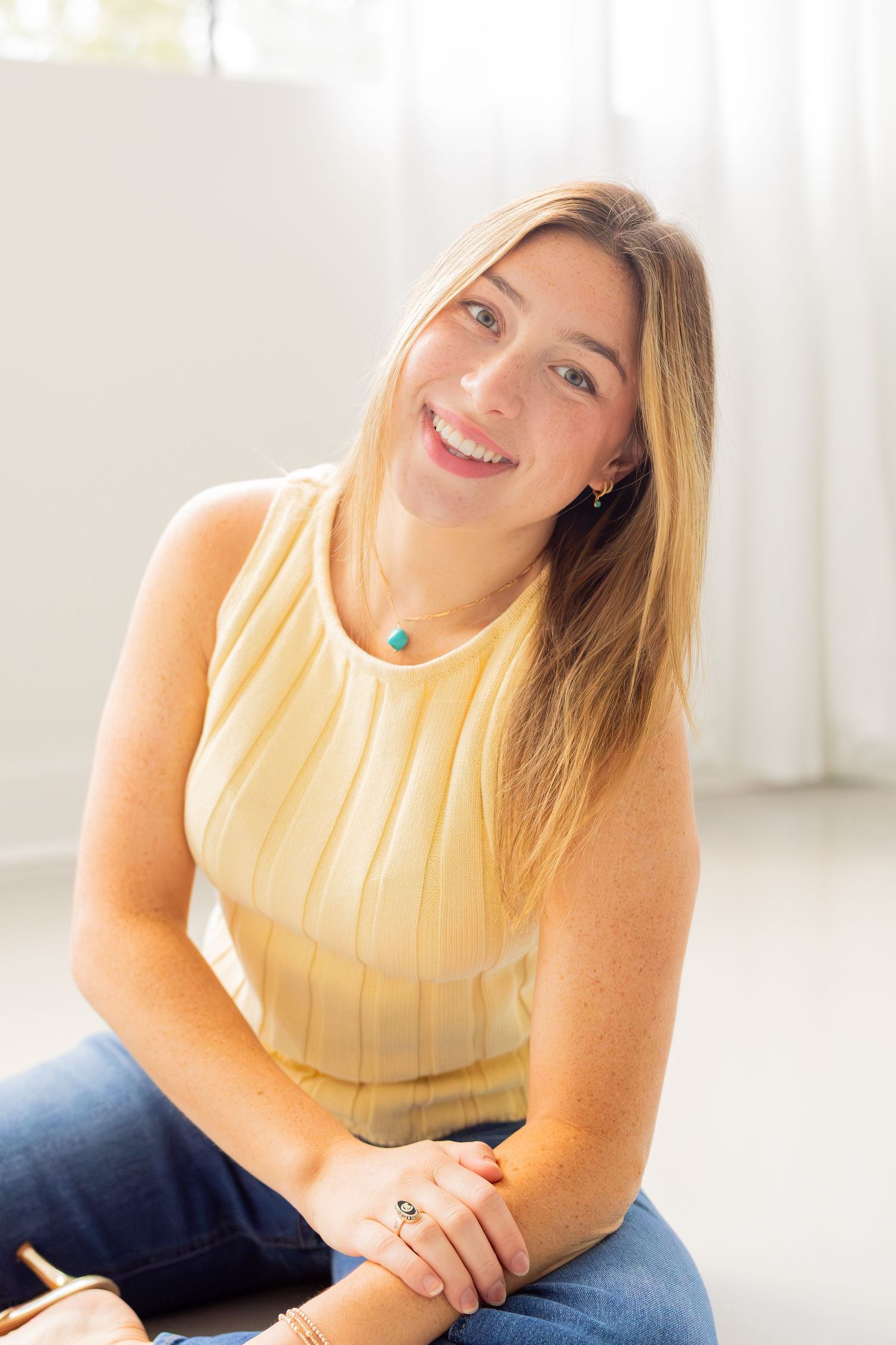 Smiling young woman with long blonde hair wearing a yellow sleeveless top and jeans, sitting relaxed indoors.