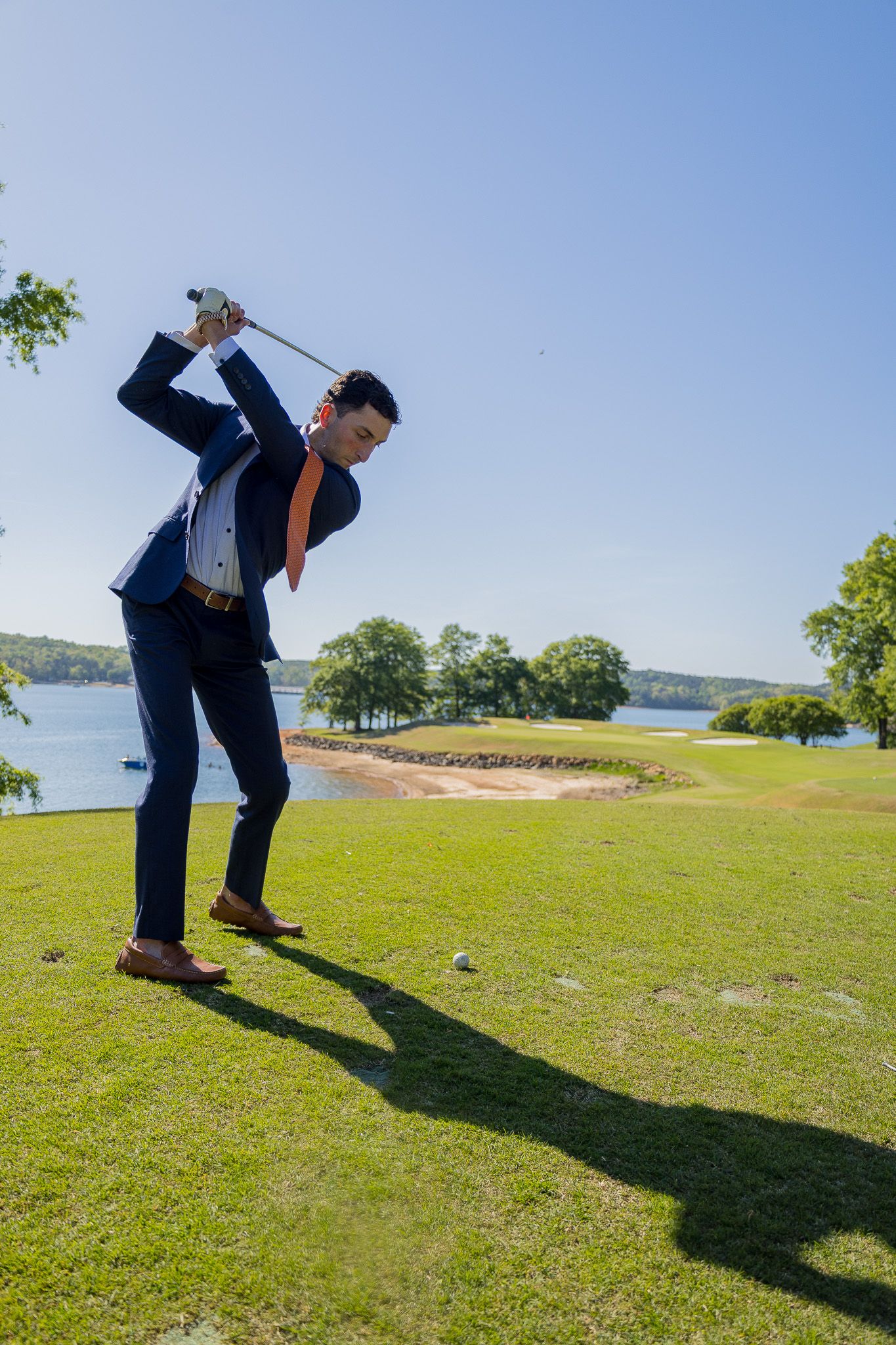 Man in navy suit and orange tie swinging a golf club on a lush green course beside a scenic lake.