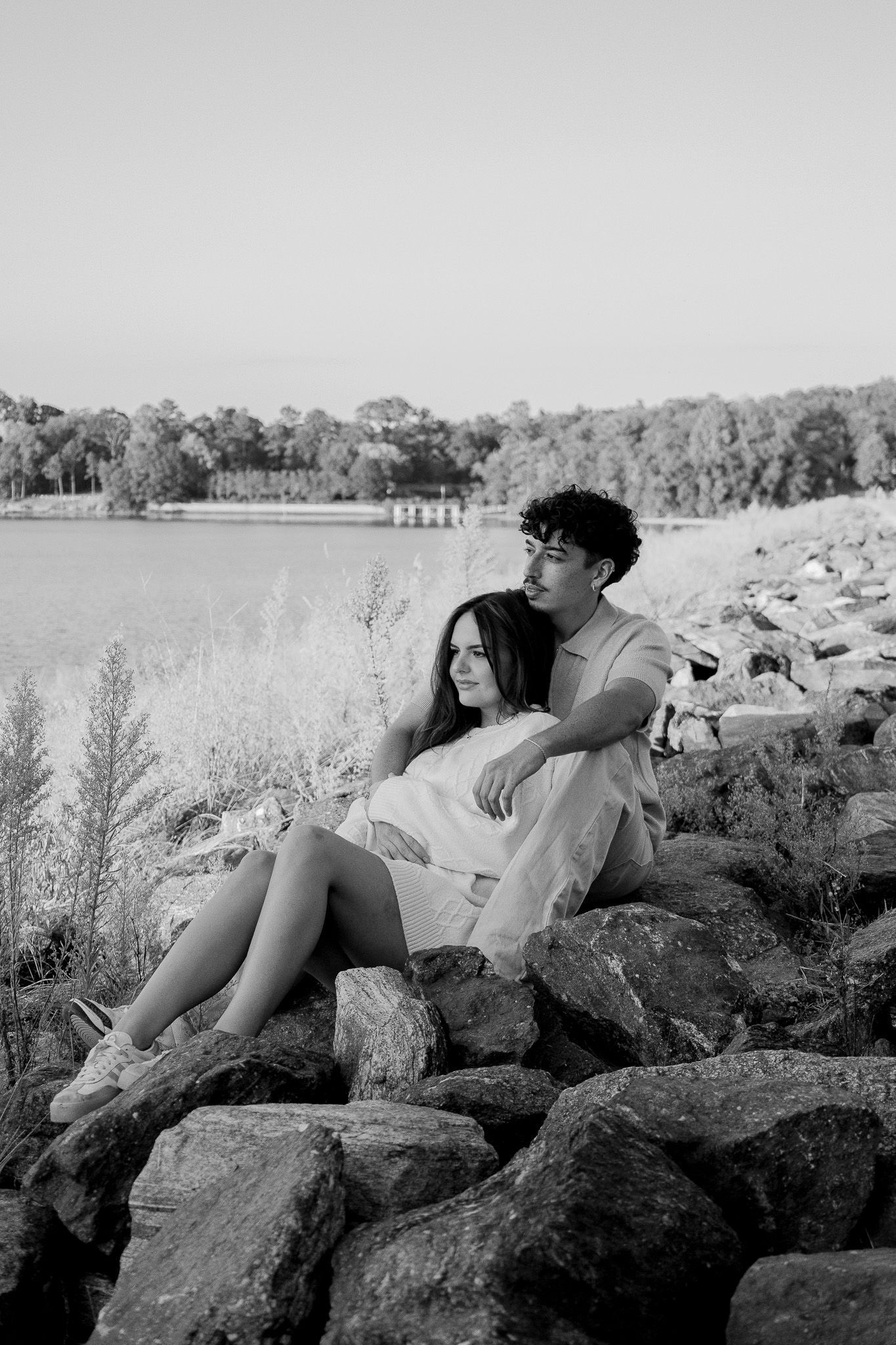 Black and white photo of a couple sitting on rocks by a lake, the woman reclining against the man as they gaze into the distance.