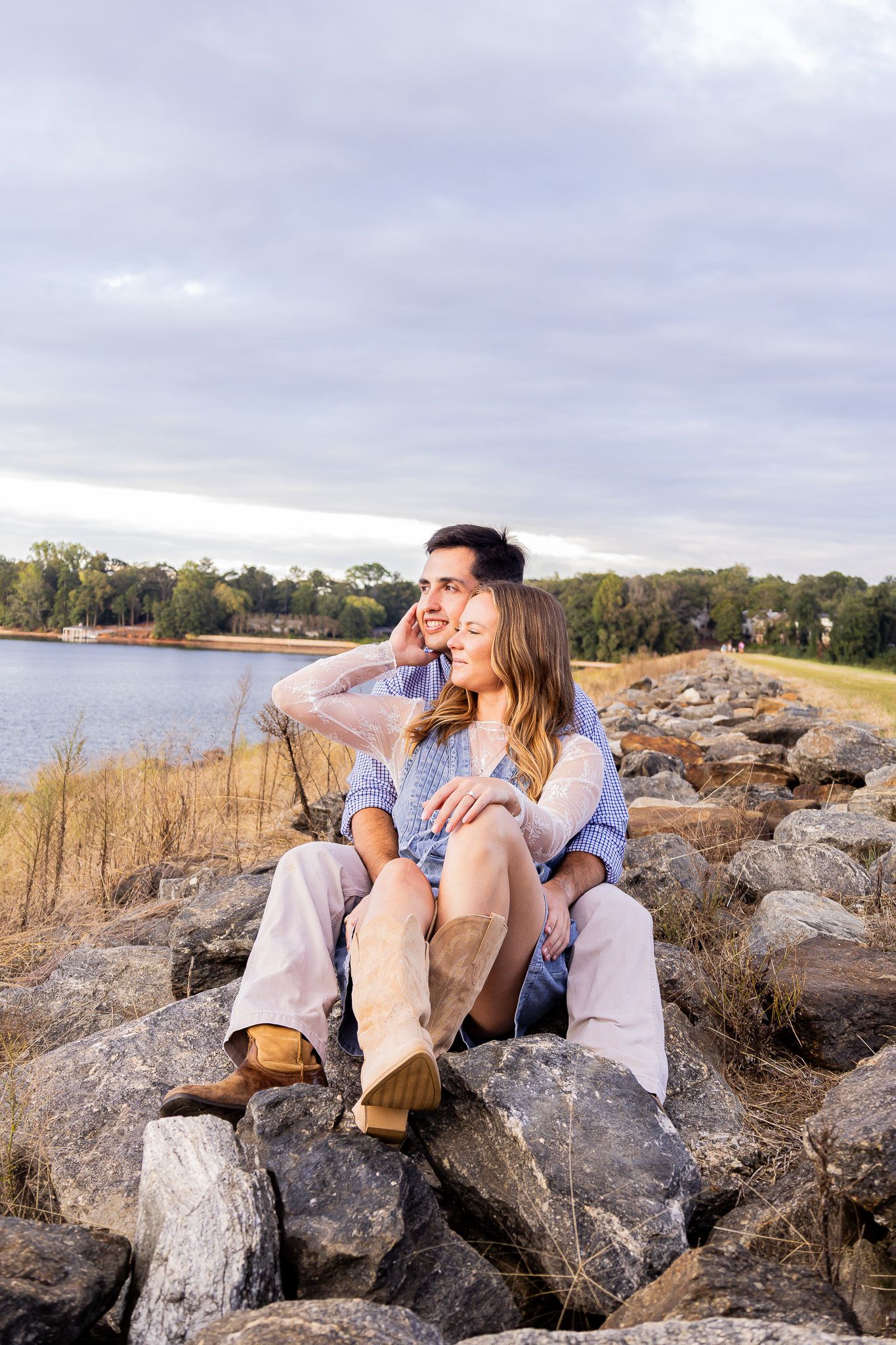A couple sits on rocky shoreline, woman leaning back on man, both smiling and gazing at a serene lake under cloudy skies.