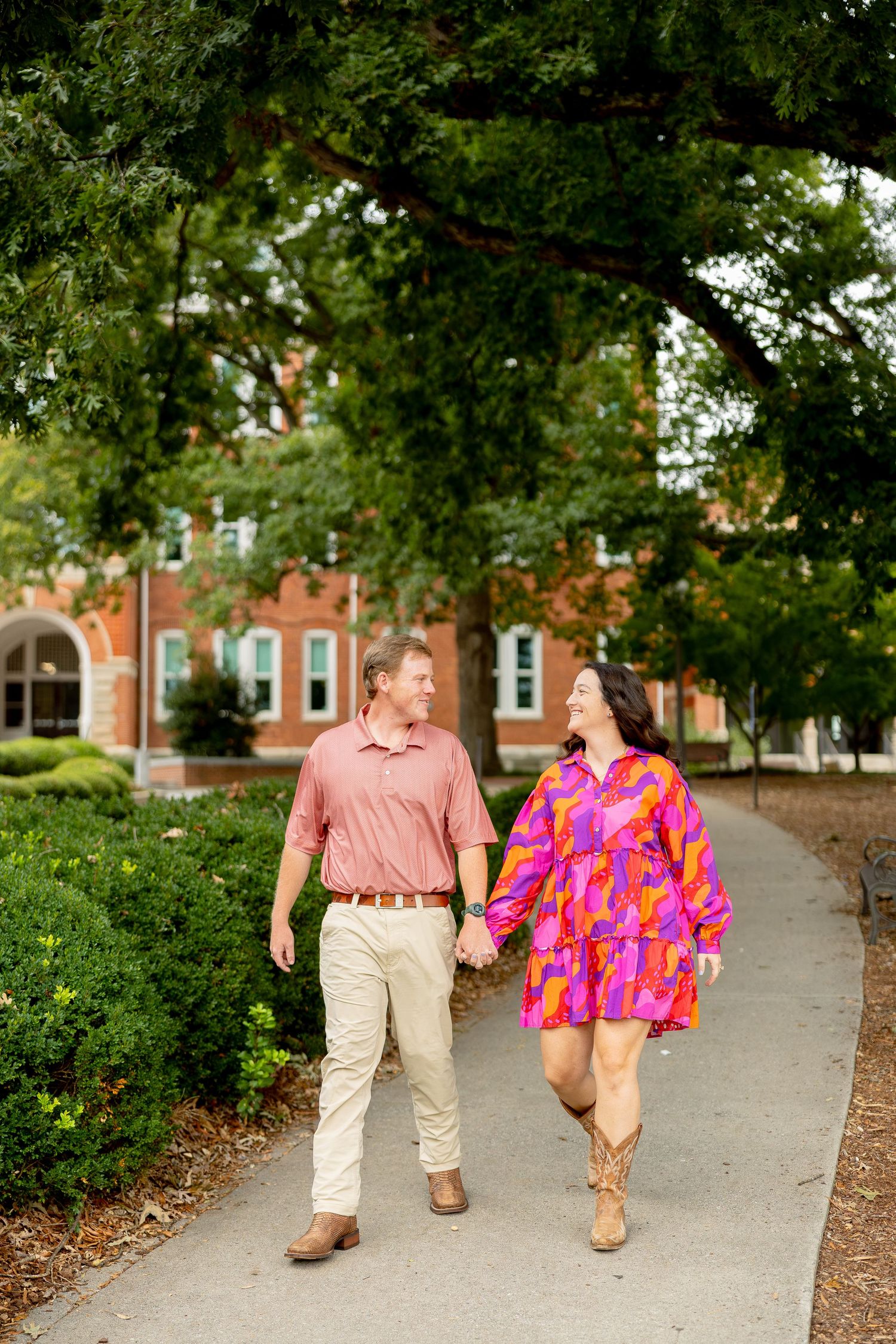 Couple holding hands walking on a path under trees near a brick building, wearing pink and colorful outfits.