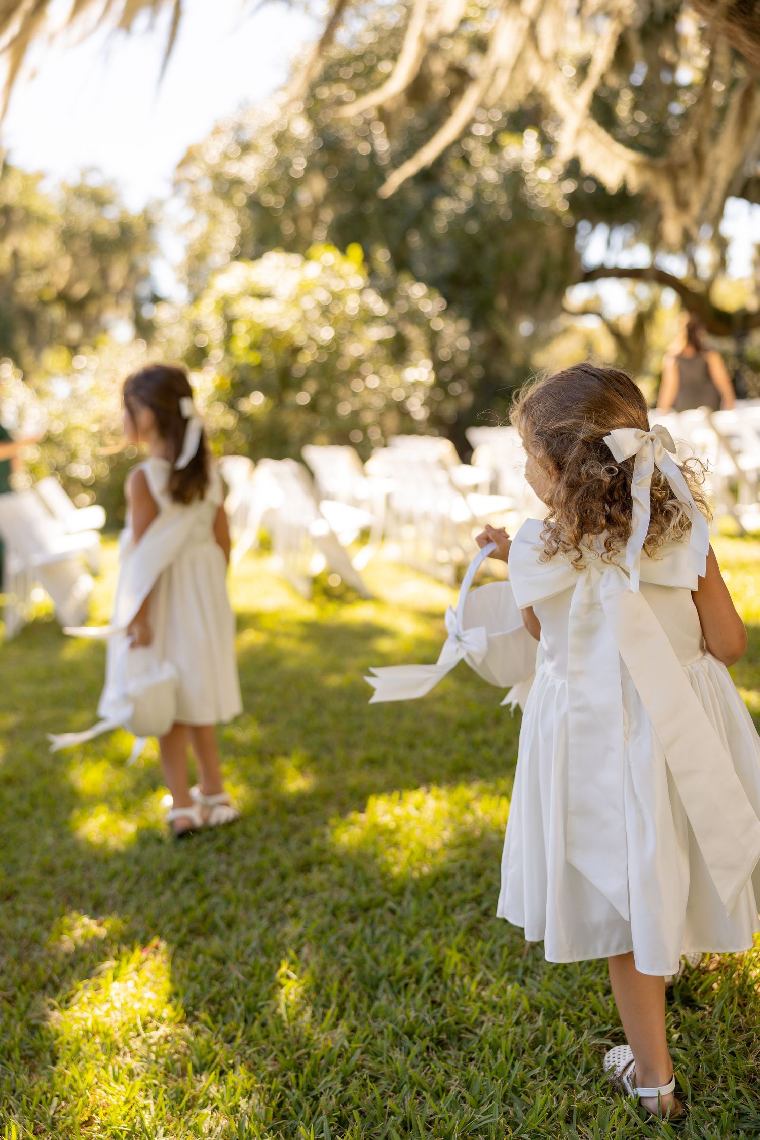 Two flower girls in white dresses with large bows walk across green grass at an outdoor wedding ceremony.