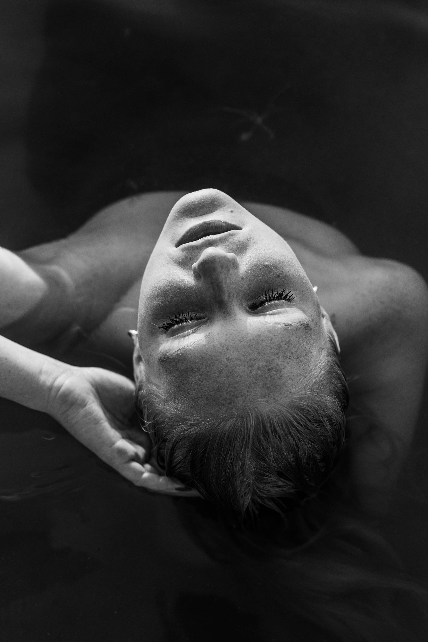 Black and white photo of a person floating in dark water with eyes closed, hair spread out, in a serene pose.
