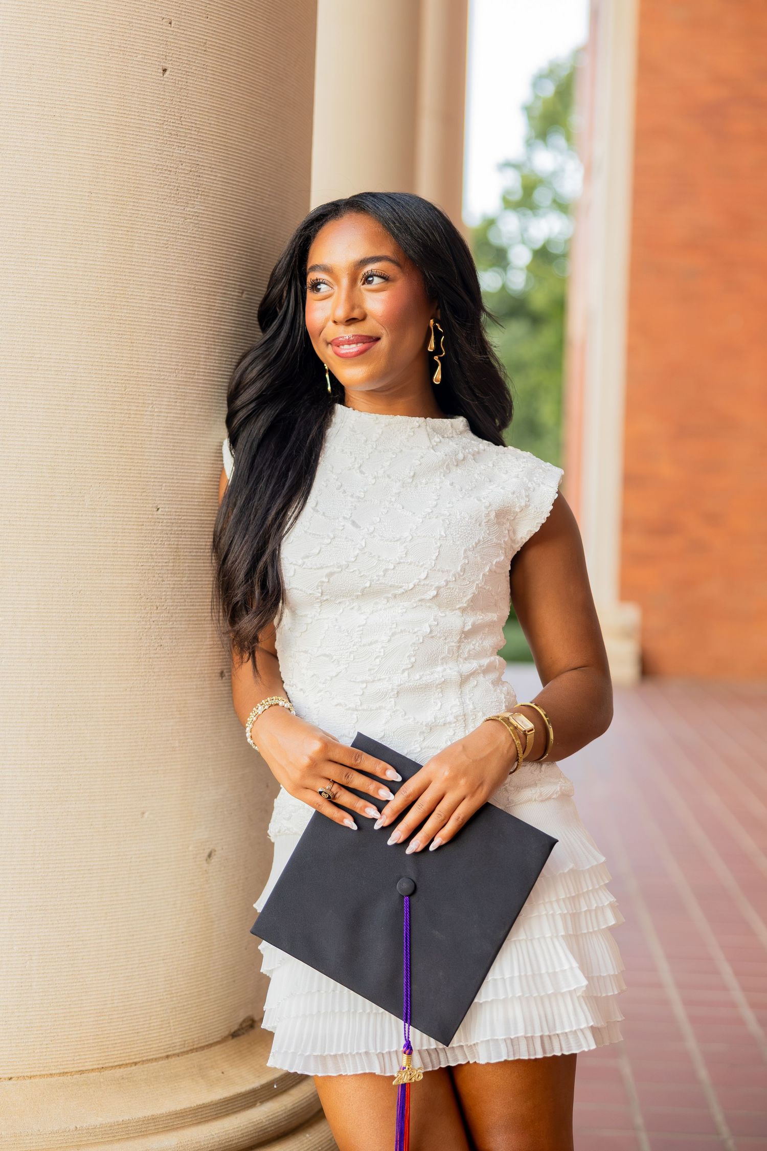 Young woman in white dress holding graduation cap, leaning against column, smiling outdoors.