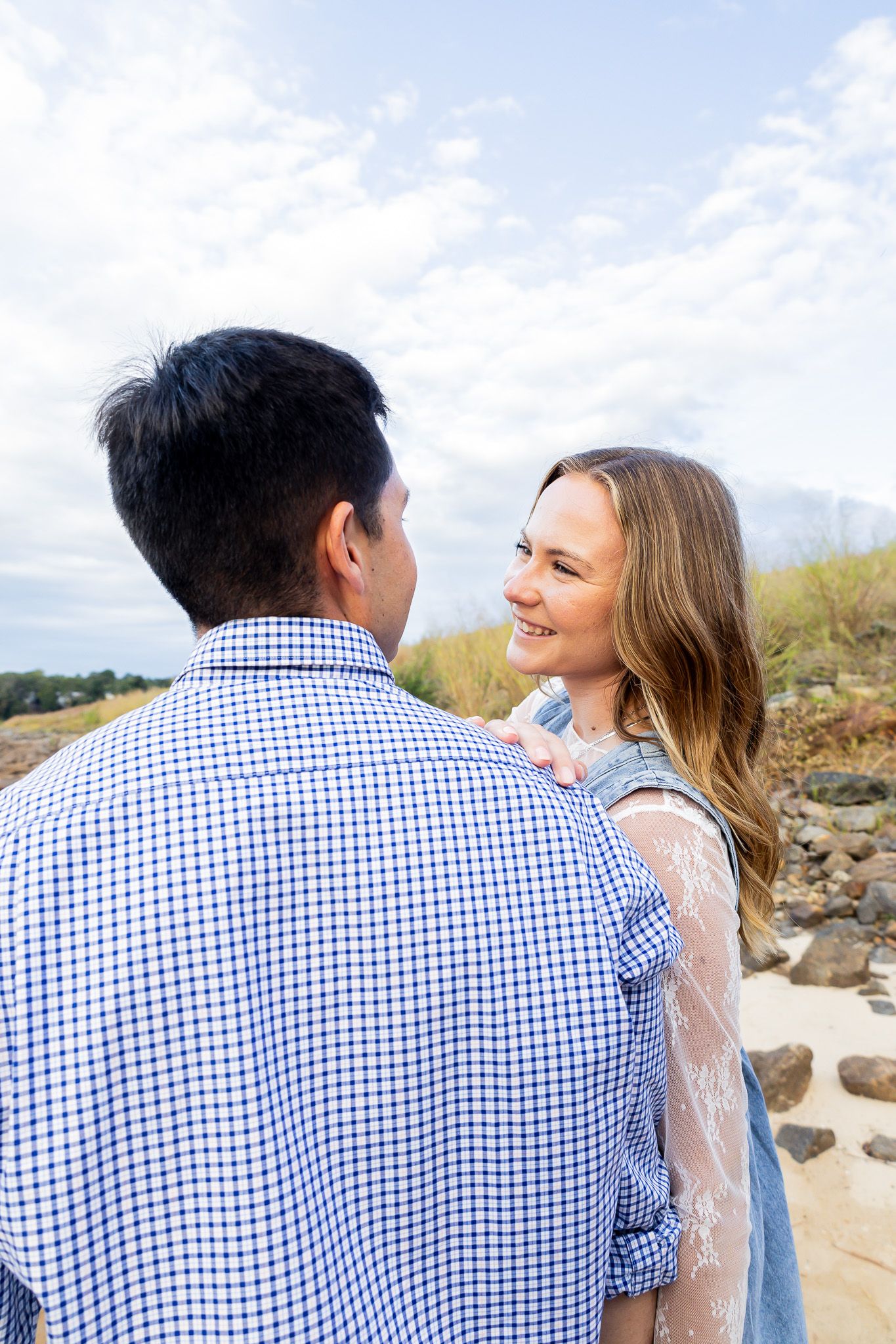 A smiling young woman gazes at a man in a blue checkered shirt outdoors near rocky terrain under a cloudy sky.