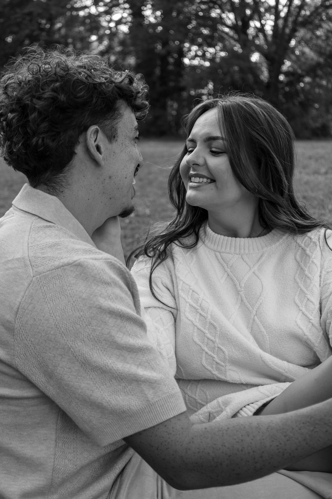 Black and white photo of a smiling couple embracing outdoors, sharing a tender moment in a park setting.
