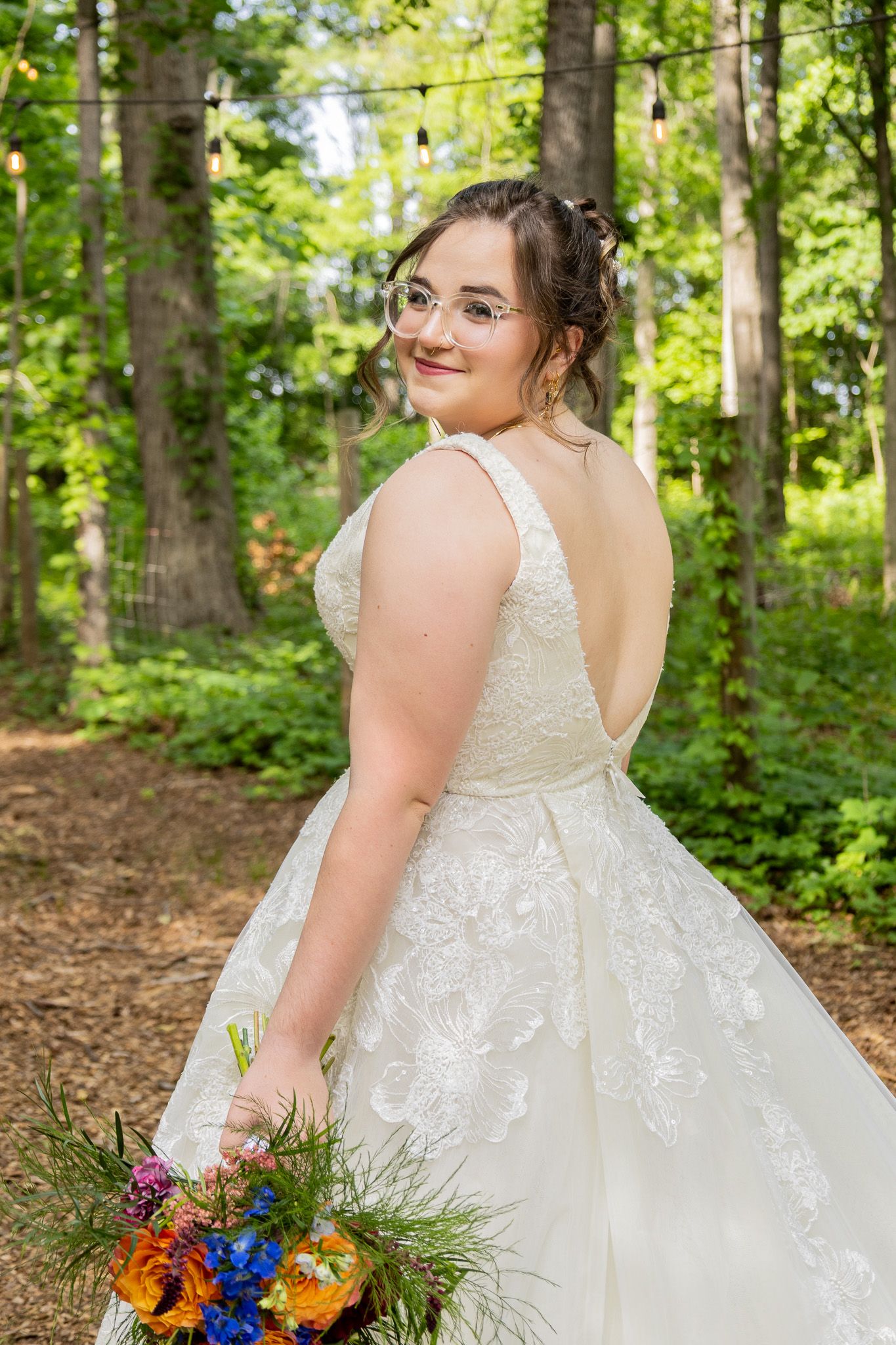 Bride in backless lace wedding gown holds colorful bouquet, smiling over shoulder in lush forest setting.